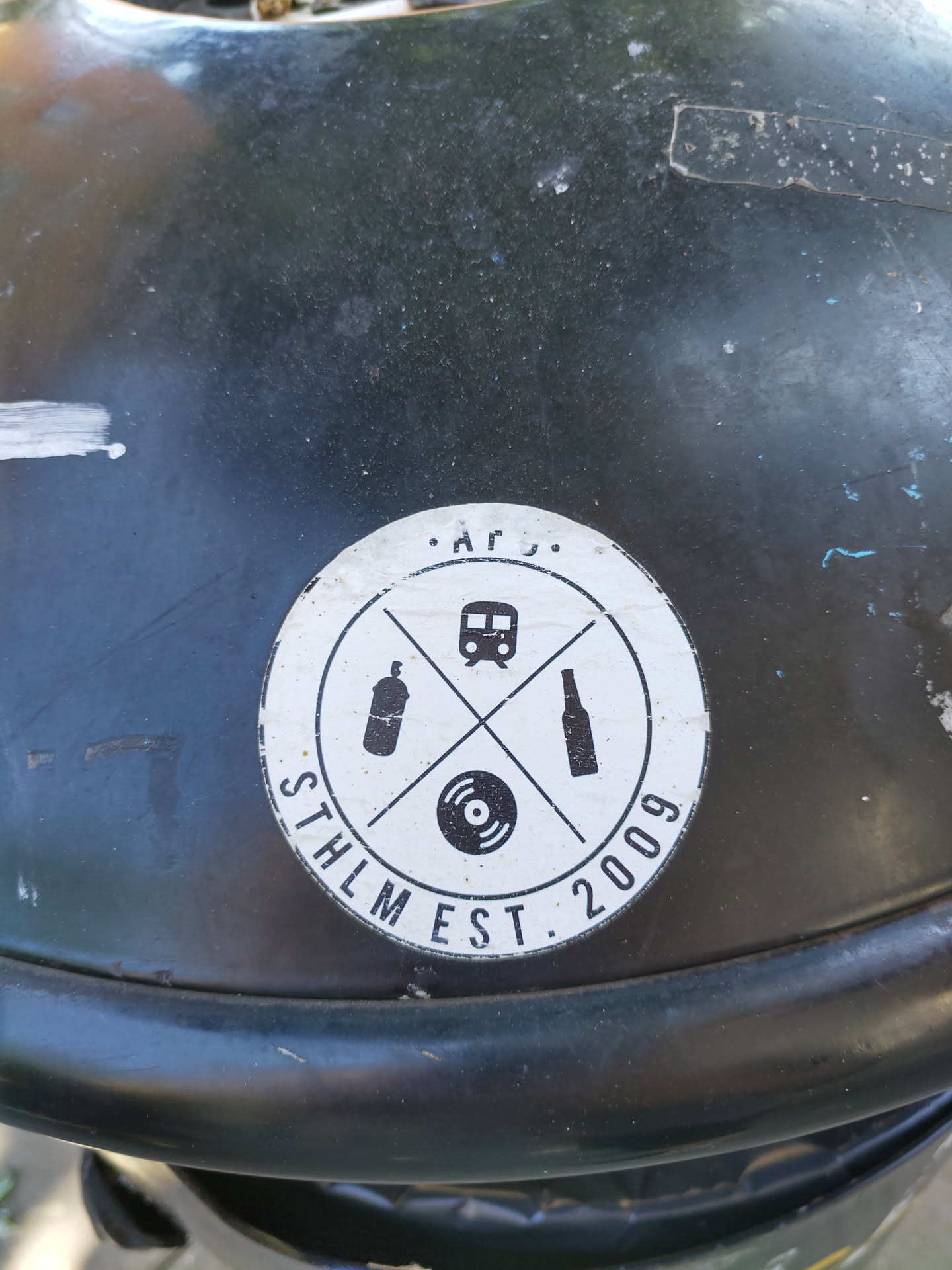 A round black and white sticker with a logo. The logo features a train, a spray can, a bottle and a vinyl record crossed in an 'x' shape. The text 'STHLM EST. 2009' is visible.  It appears to be a logo for a Stockholm-based establishment founded in 2009, perhaps a bar, club or record store.