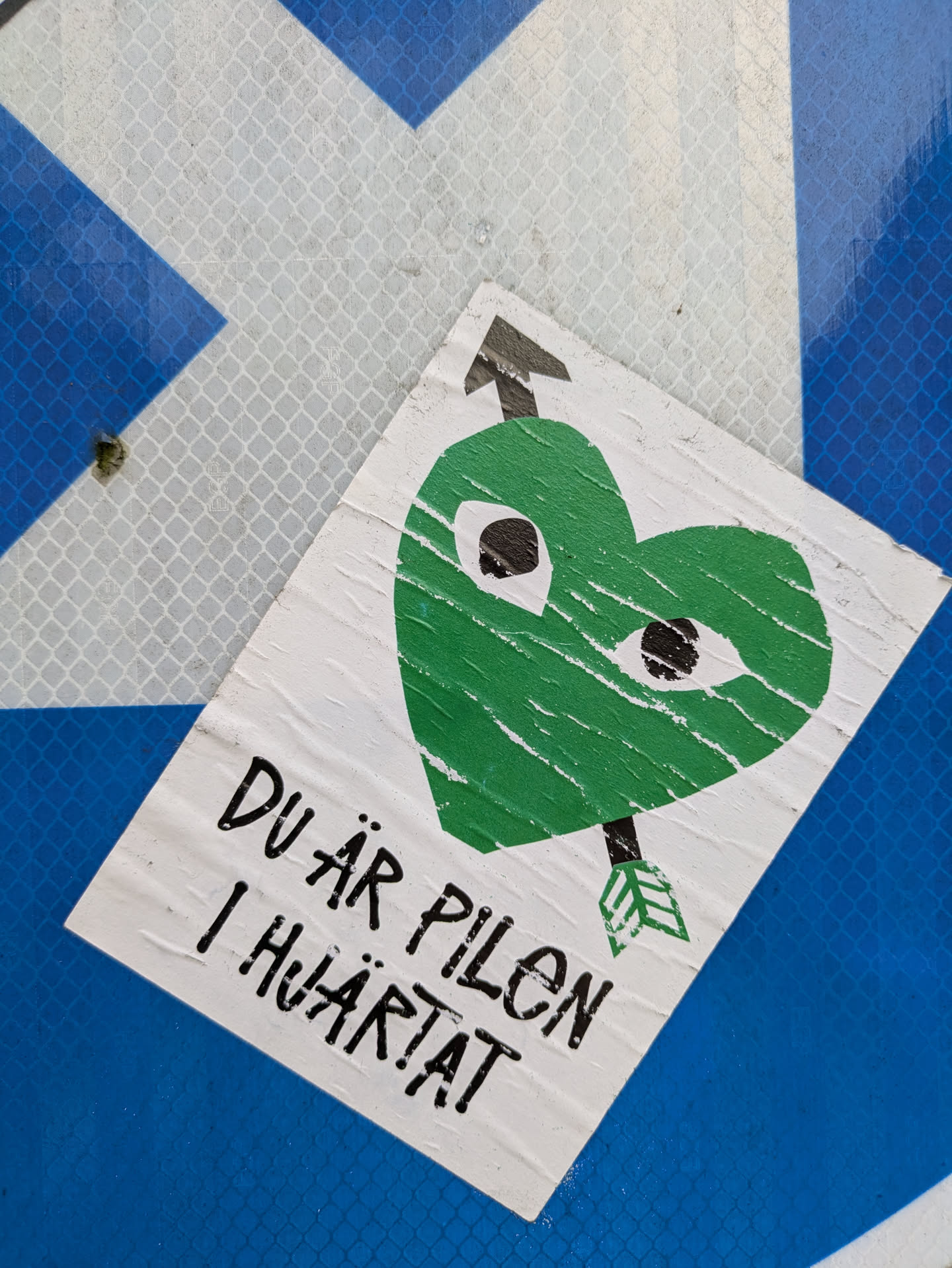 A sticker featuring a green heart with eyes and an arrow piercing it. The text below translates to "You are the arrow in the heart." The sticker shows some signs of wear and tear from being stuck on a surface.