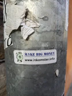 A sticker featuring a cartoon money bag and the text 'MAKE BIG MONEY' with a website address underneath.  It's adhered to a weathered metal pole.