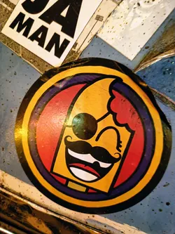 This round sticker features a cartoon-like character with a yellow face, red hair, a black mustache, and sunglasses. It is winking and has a cheerful expression. The character is surrounded by concentric circles of purple, red, and gold.