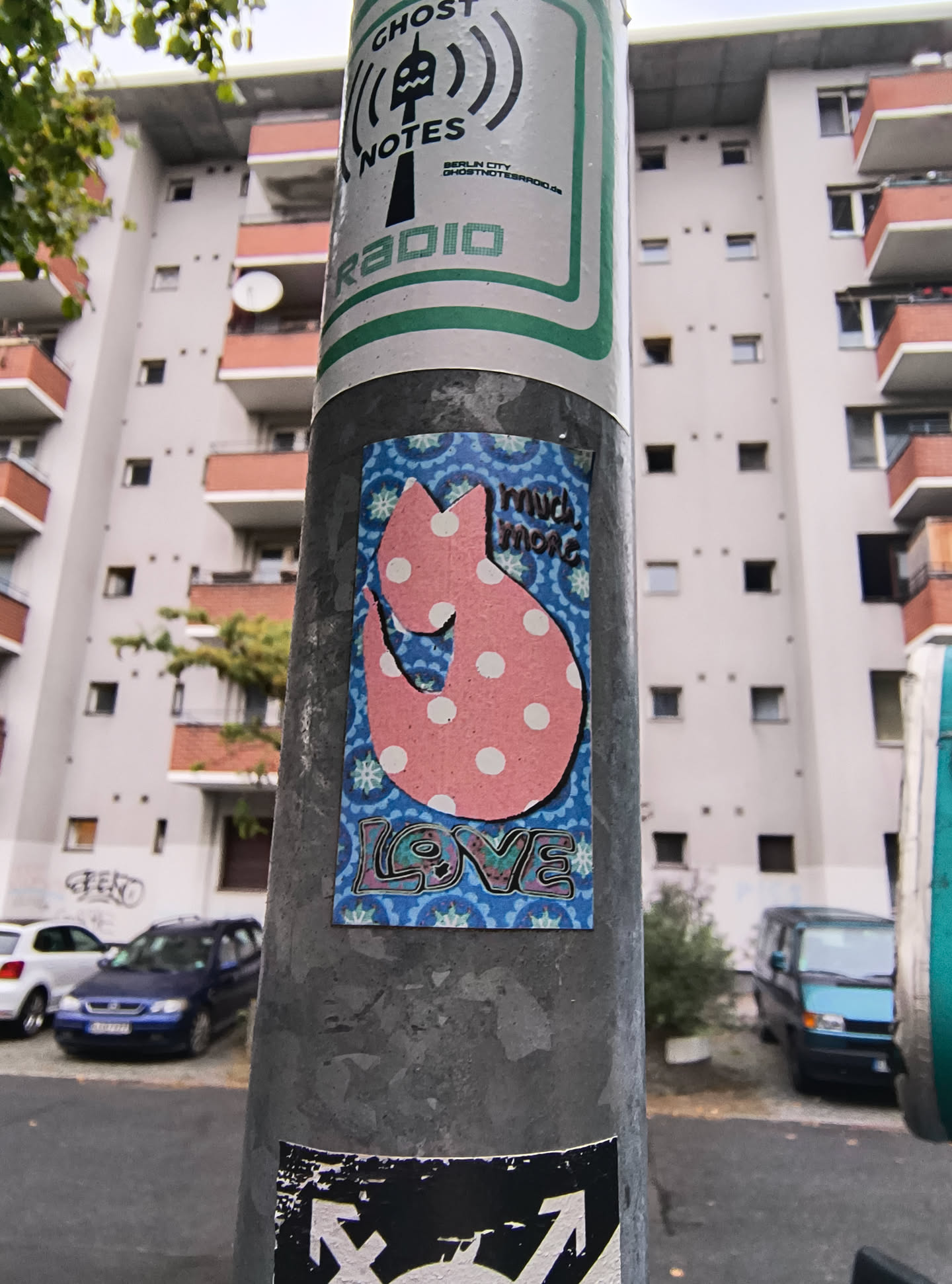 The sticker is predominantly blue with a repeating wave-like pattern. Centered on the sticker is a stylized pink cat silhouette filled with white polka dots. The cat has a curved tail and appears to be in a playful pose. Above the cat, in a darker pink, are the words 'much more'. Below the cat, in a colorful, graffiti-style font with pink, blue, and green hues, is the word 'LOVE'. The overall aesthetic is cheerful and whimsical.