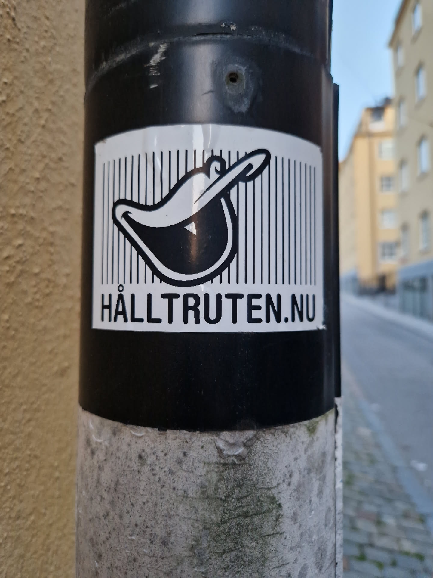Black and white sticker with a cartoon-like image of a hat and the website address Hälltruten.nu. The sticker is affixed to a grey pole.