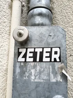 A black and white sticker with the word ZETER in bold, uppercase letters is affixed to a weathered metal surface. The sticker is slightly worn at the edges.  The background is a gray metal box, possibly an electrical box or similar utility component, attached to a building's exterior wall.