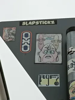 A sticker featuring a drawing of a three-headed character with a single eye in the center. The style is cartoonish and slightly grotesque. The sticker is part of a collection by Slapstickz, as indicated by another sticker on the same surface.