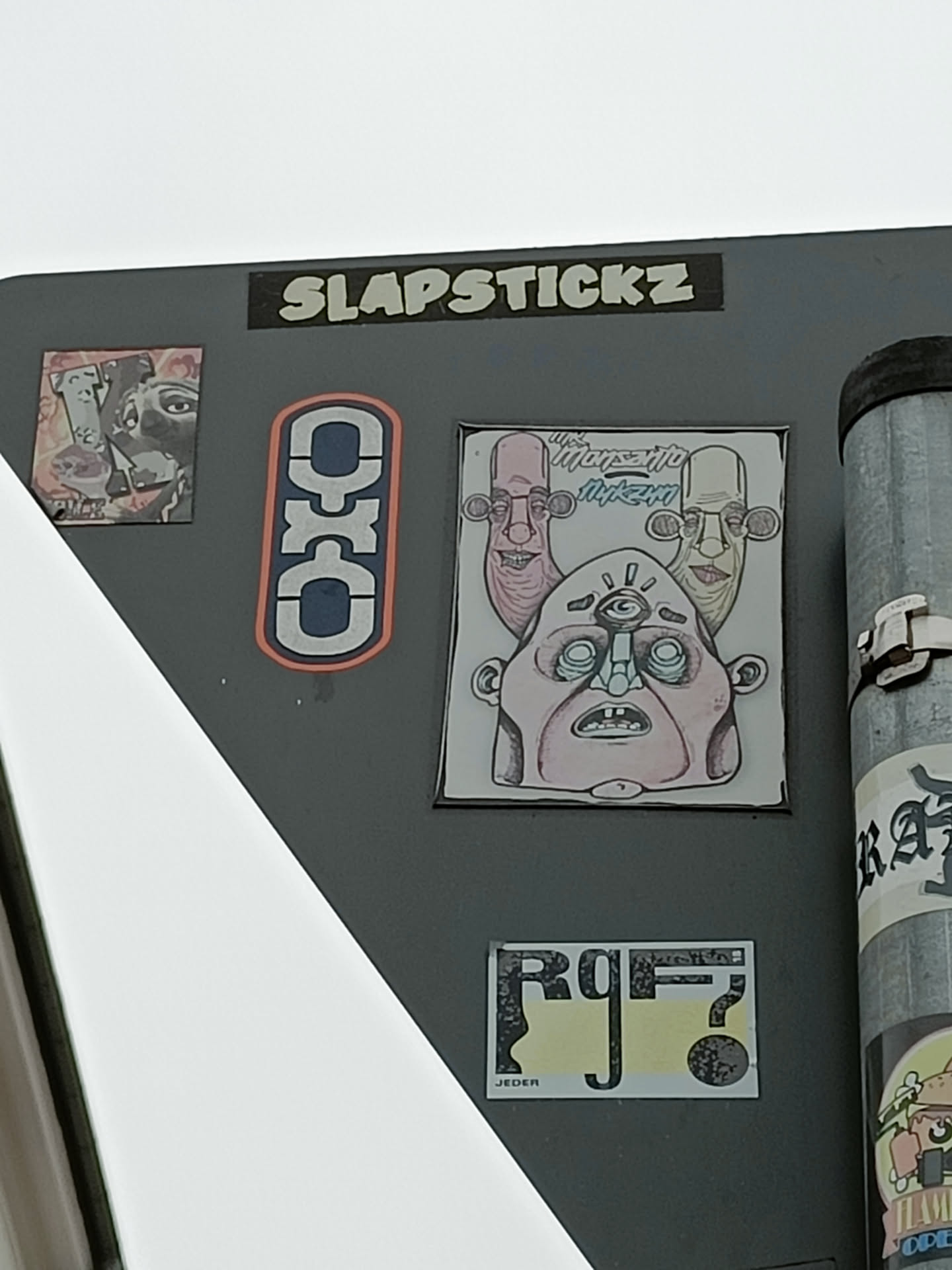 A sticker featuring a drawing of a three-headed character with a single eye in the center. The style is cartoonish and slightly grotesque. The sticker is part of a collection by Slapstickz, as indicated by another sticker on the same surface.