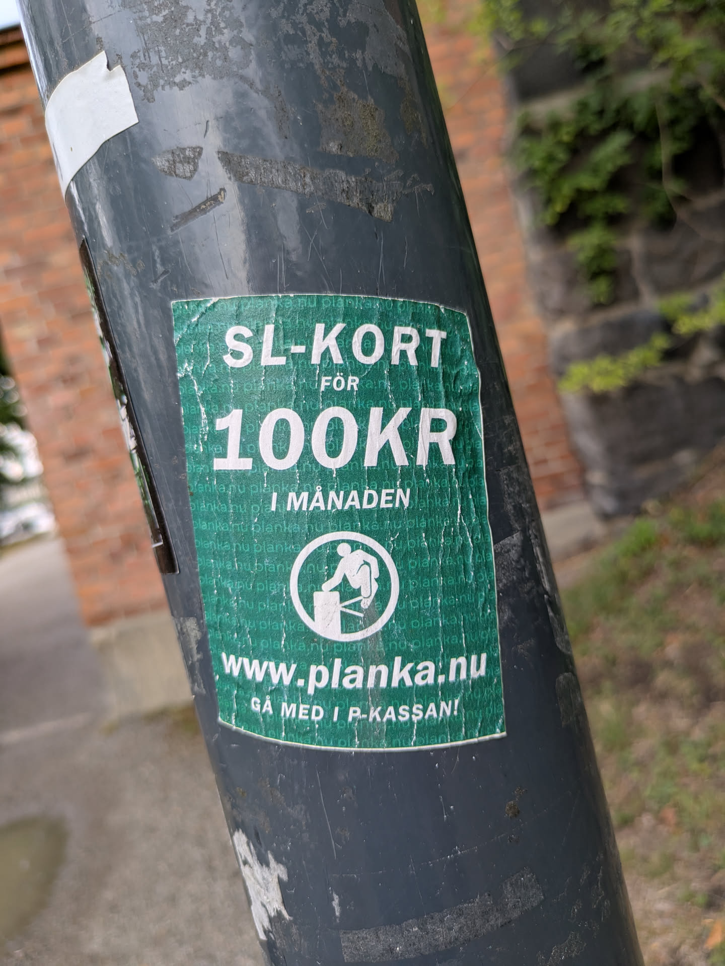 The sticker is a rectangular, dark teal or forest green decal affixed to a dark grey metal pole. It shows significant signs of wear, including creases, fading, and peeling edges. The main text, in white sans-serif font, reads "SL-KORT FÖR" at the top, followed by "100KR" in a larger font, and then "I MÅNADEN". Below this, a white circular logo depicts a stylized figure jumping over a turnstile or barrier. The background of the sticker features a subtle, repeating pattern of the word "planka.nu". At the bottom, in white, is "www.planka.nu" and beneath that, "GÅ MED I P-KASSAN!".
