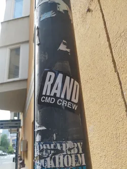 A black and white sticker with the text "RAND CMD CREW" is affixed to a pole. The sticker shows some wear. Other stickers are visible on the pole, some are damaged.