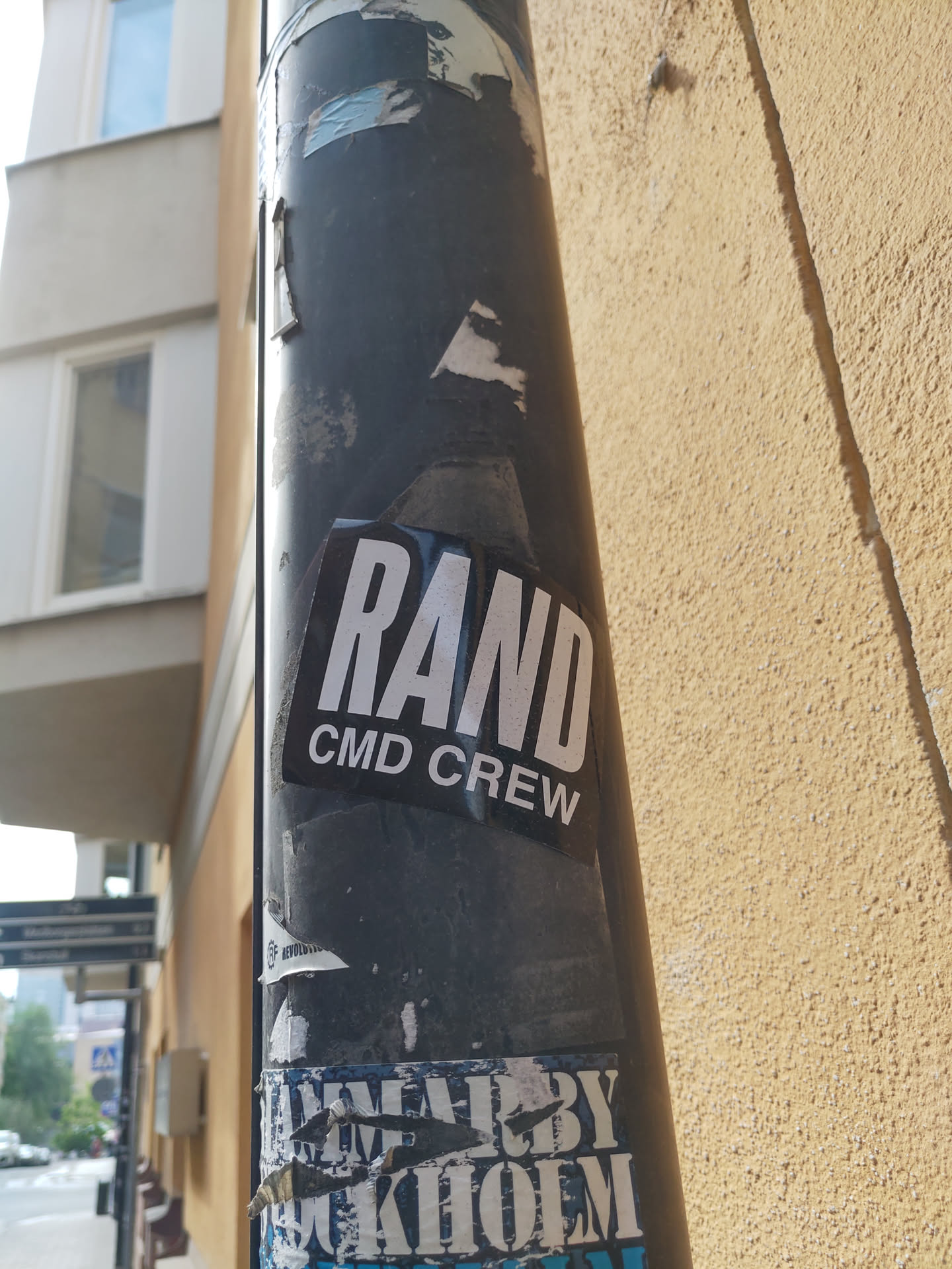 A black and white sticker with the text "RAND CMD CREW" is affixed to a pole. The sticker shows some wear. Other stickers are visible on the pole, some are damaged.