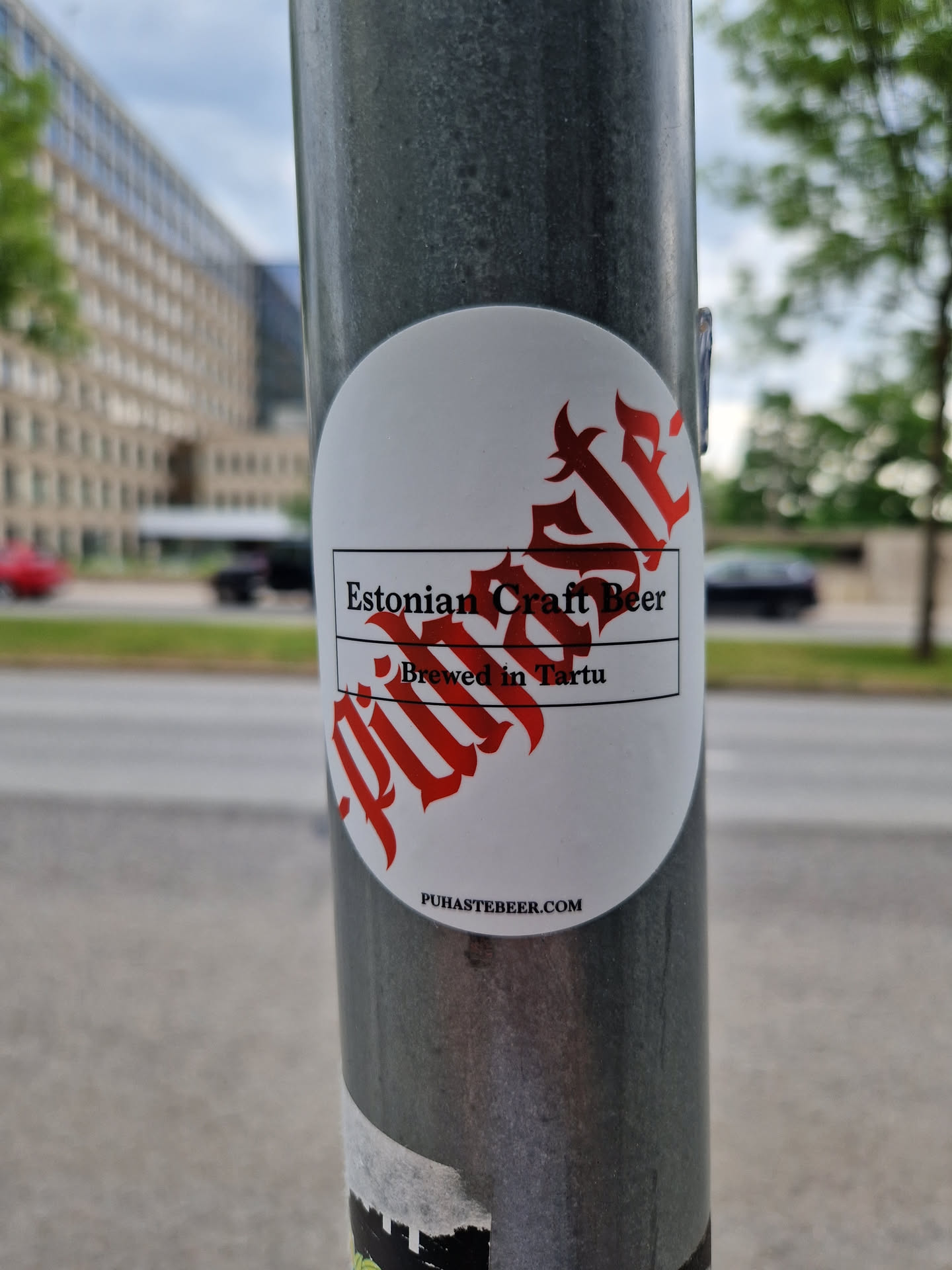 A round white sticker with red text on a gray pole.  The sticker advertises Estonian Craft Beer brewed in Tartu. A website address is also visible.