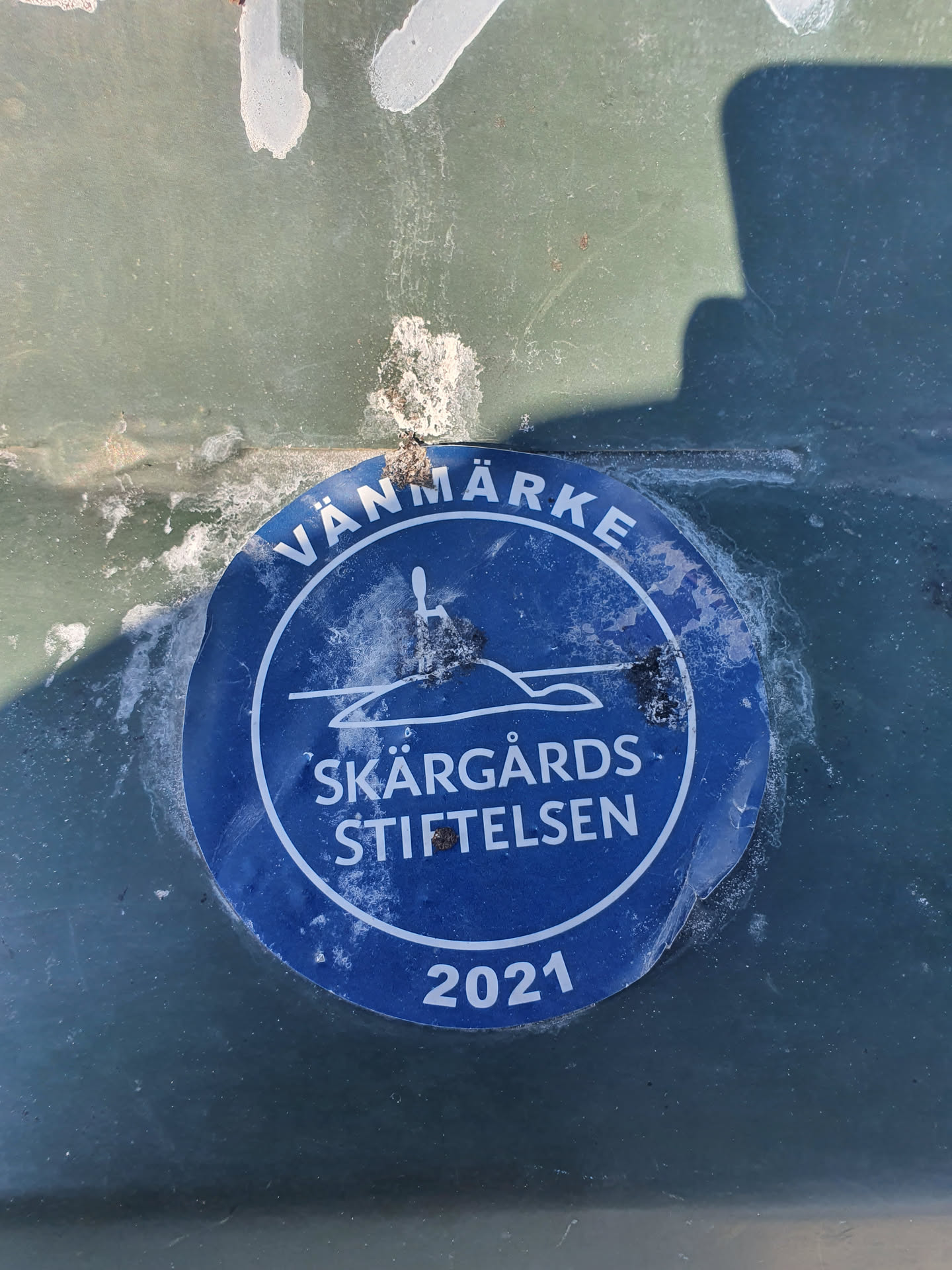A blue round sticker with the text "Vänmarke Skärgårds Stiftelsen 2021" and a picture of an island with a lighthouse.  The sticker appears to be weathered.