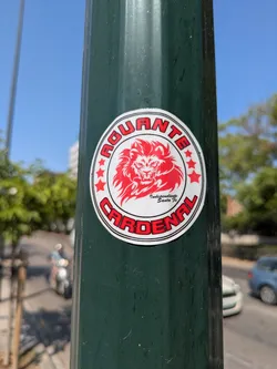A round sticker is affixed to a green pole. The sticker has a white background with a red border. Around the top border of the sticker, in red letters, is the word "AGUANTE." At the bottom border of the sticker is the word "CARDENAL," also in red letters. In the center of the sticker is a stylized red lion's head. Below the lion's head is the text "Independiente Santa Fe". There are also five red stars along the side.
