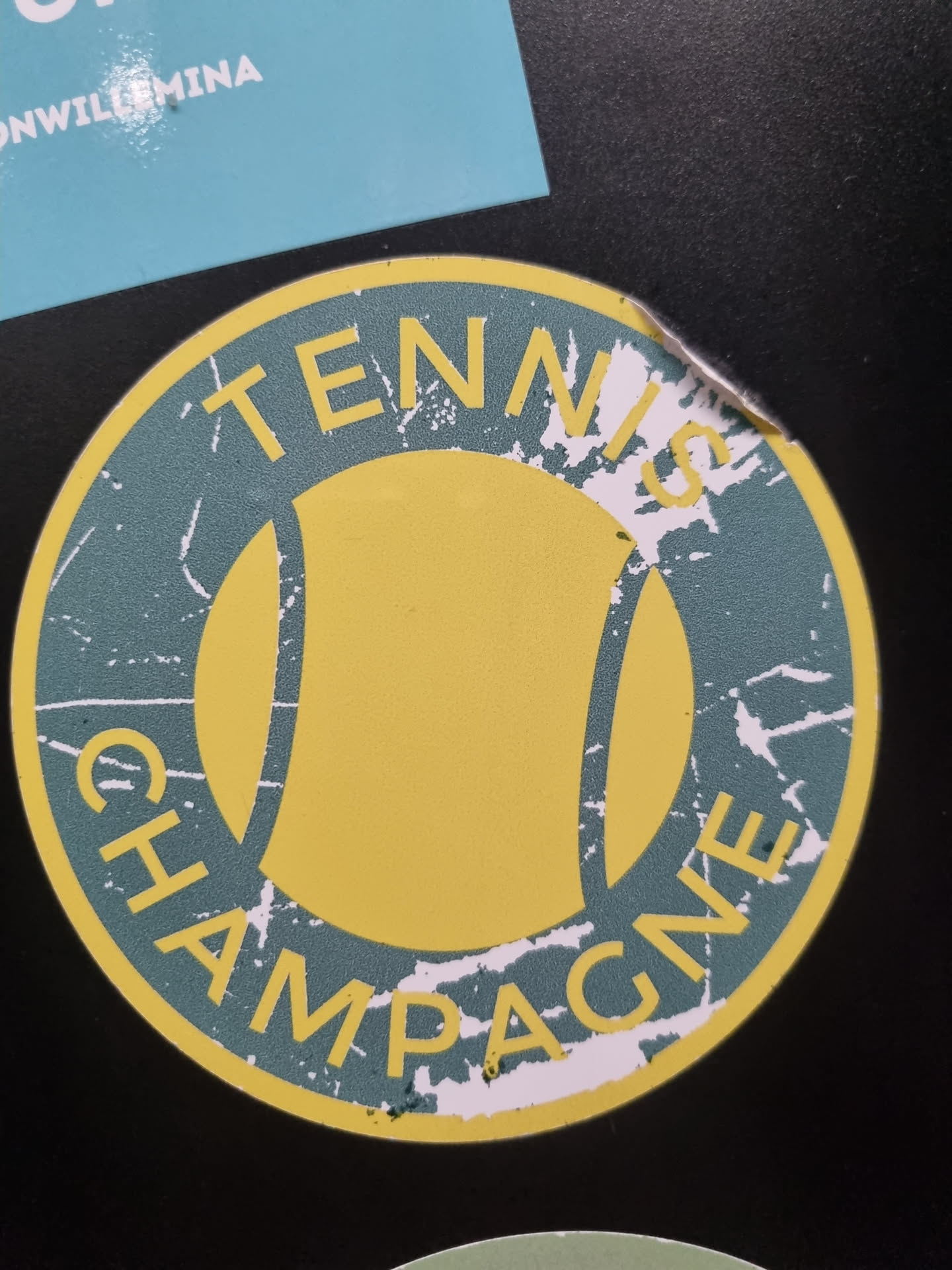 Round sticker with a distressed design. It features a tennis ball in yellow in the center with the words "Tennis" at the top and "Champagne" at the bottom, all in a dark teal color. The sticker shows some wear and tear around the edges.