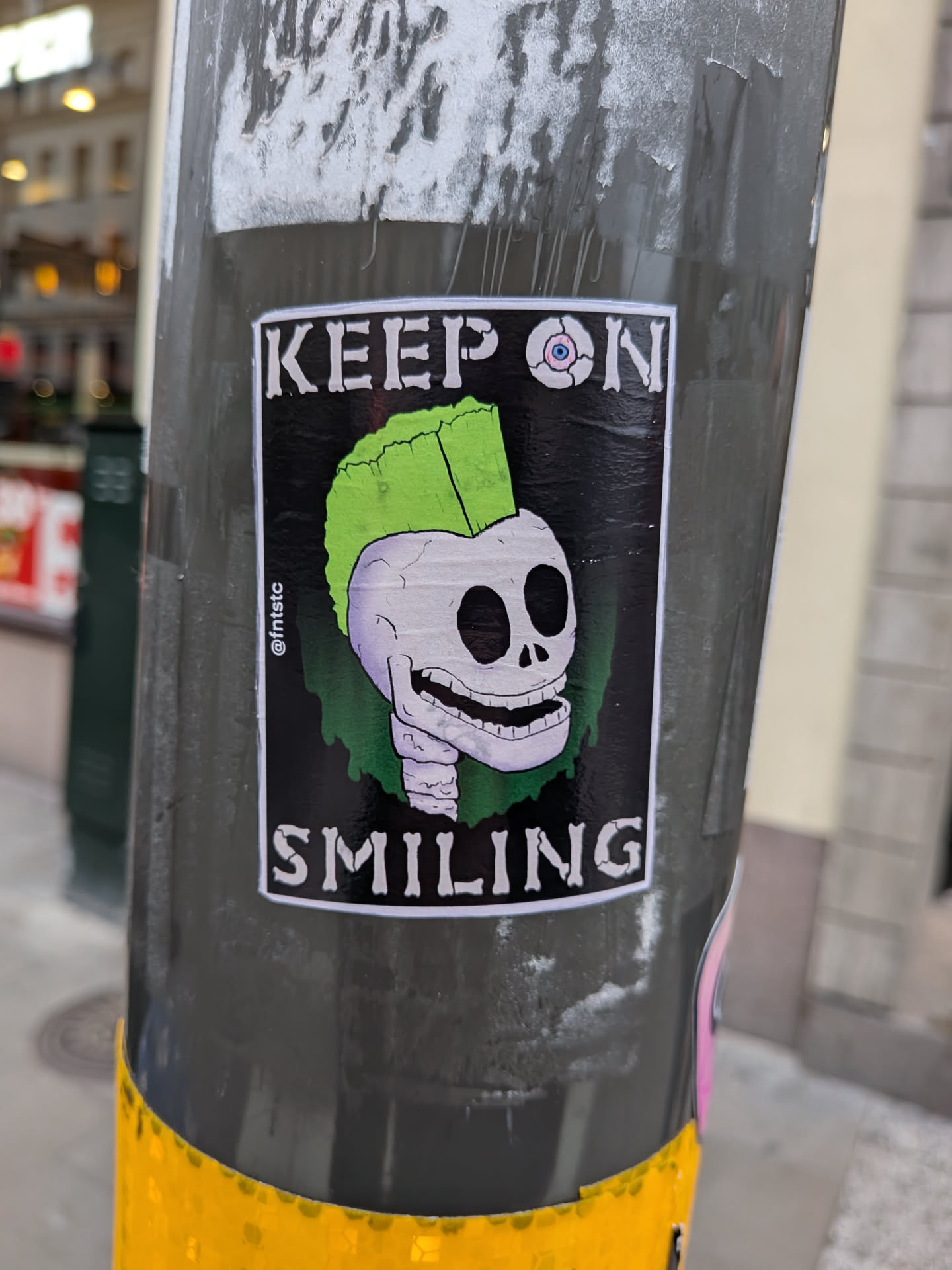 The sticker features a cartoonish skull with a bright green mohawk, a wide, toothy grin, and empty black eye sockets. One eye socket contains a stylized eyeball with a pink iris and blue pupil. The skull is depicted with a light purple hue and is outlined in black. The background is black, with green dripping elements behind the skull. The text 'KEEP ON' is at the top and 'SMILING' is at the bottom, both in white, stylized, bone-like lettering with a black outline. A small '@fntstc' handle is visible vertically on the left side of the sticker.