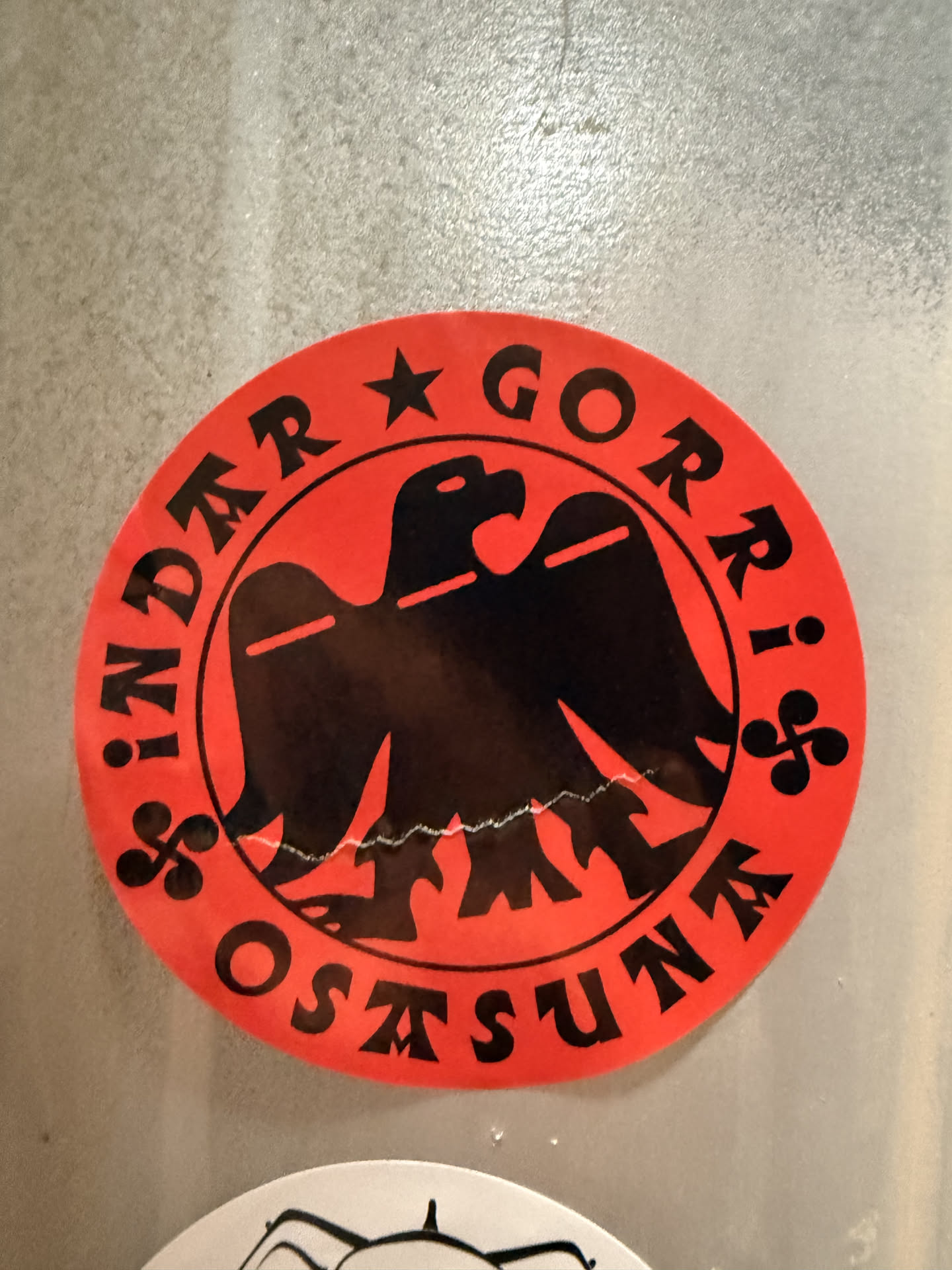 A circular sticker with a bright red background and black elements. At its center is a stylized black eagle, facing left with its wings spread. The eagle has a sharp beak and a single eye, and its lower body features a jagged, wave-like or mountainous outline. Encircling the eagle is black text: 'INDER ★ GORRI' along the top arc and 'OSTASUNA' along the bottom arc. A black five-pointed star separates 'INDER' and 'GORRI'. Two black Lauburu symbols (a traditional Basque cross with curved arms) are positioned on either side of the eagle, separating 'GORRI' from 'OSTASUNA'. The sticker shows minor signs of wear, including a slight crease or imperfection near the bottom edge of the eagle.