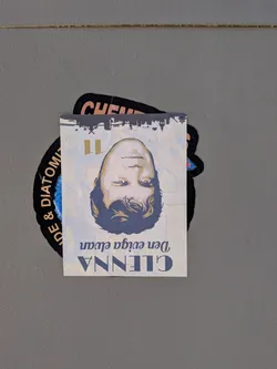 The sticker is a rectangular piece of paper with a portrait of a person's face, oriented upside down. The face is rendered in a stylized, illustrative manner with bold lines and a limited color palette of skin tones, dark blue for hair and shadows, and yellow for accents. Above the face, a silhouette of a cityscape is depicted. Below the face, text is printed in a vintage-style font. The sticker is partially overlapping a larger, black, circular sticker with white text and a blue fuzzy center.