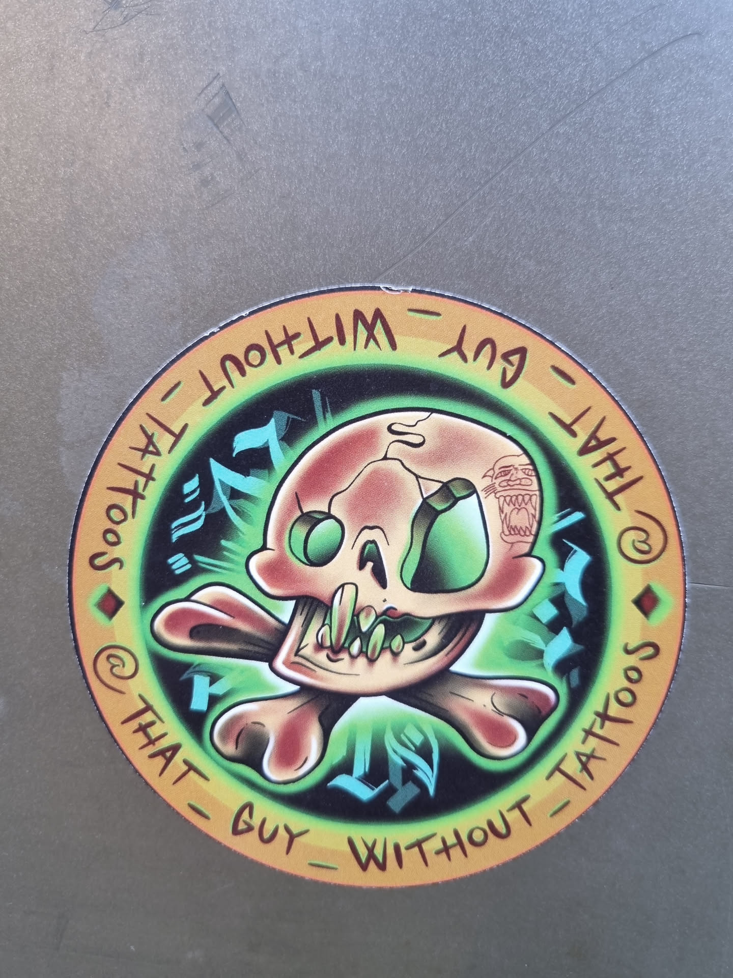 Round sticker featuring a skull and crossbones design. The skull is cartoonish and colorful with green eyes. The text around the artwork says "That Guy Without Tattoos" in a circular pattern.