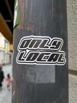 A black and white sticker with the text "Only Local" in a bold, stylized font. The sticker is affixed to a weathered gray pole.