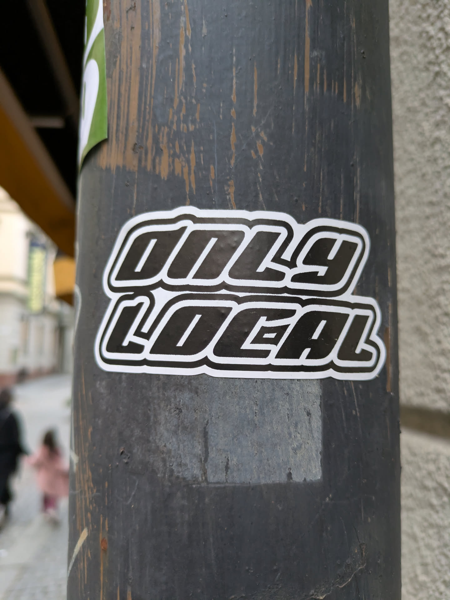 A black and white sticker with the text "Only Local" in a bold, stylized font. The sticker is affixed to a weathered gray pole.