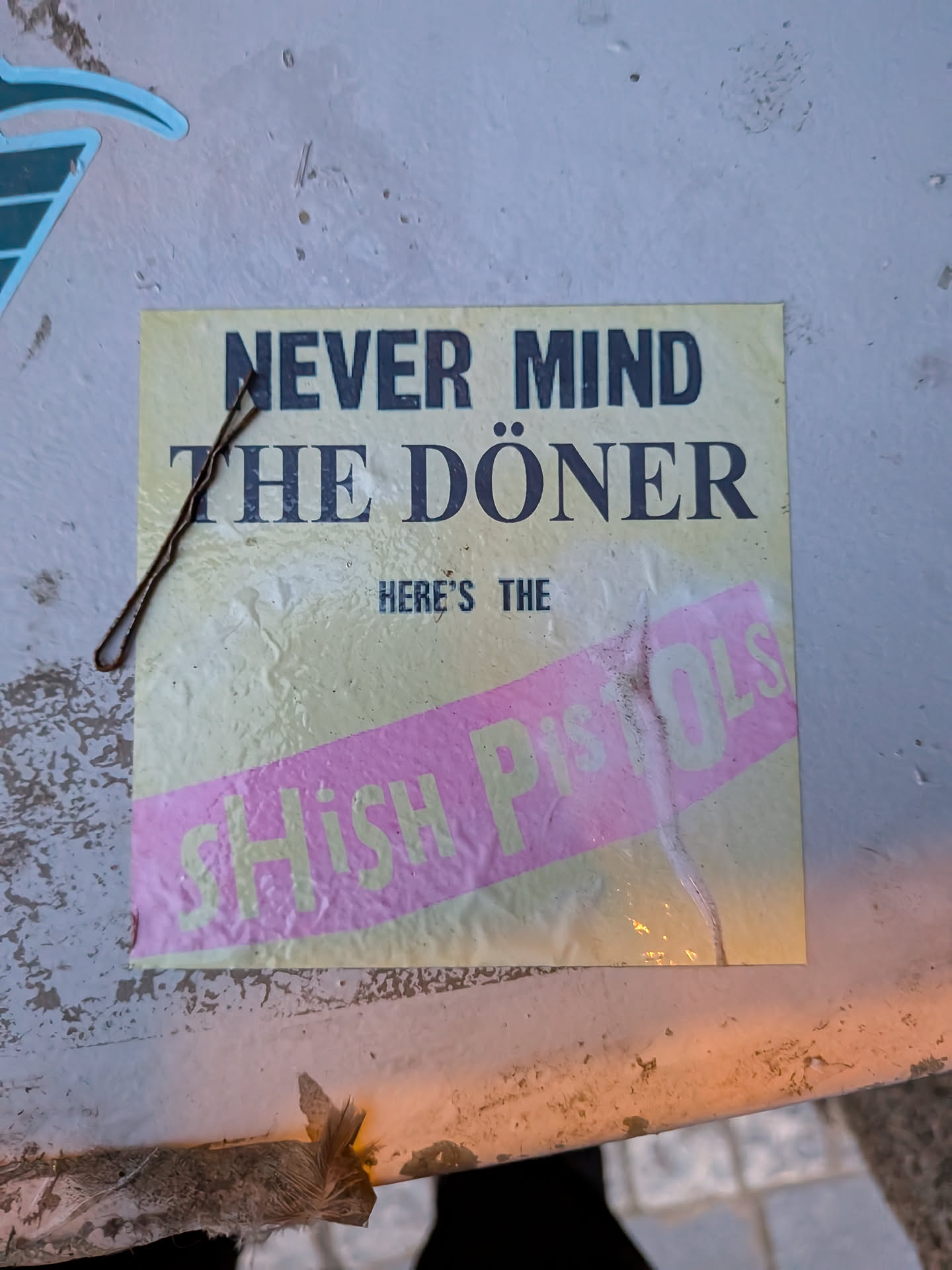 The sticker is rectangular with a pale yellow background. The top line of text in bold, dark capital letters reads 'NEVER MIND'. Below this, in slightly larger, dark capital letters with umlauts over the 'o', it reads 'THE DÖNER'. Centered below this is smaller text that reads 'HERE'S THE'. A diagonal pink band runs across the bottom half of the sticker, with the words 'Shish Pistols' printed in a lighter, almost white color with a subtle yellow outline. The sticker appears slightly worn and has some creases and a bobby pin resting on its surface.