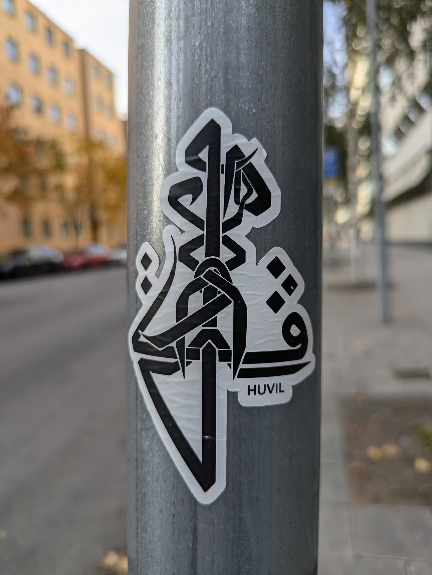 A sticker featuring a bold, black and white abstract design. The style is geometric and minimalist, with sharp lines and shapes creating an intricate pattern.  A small text 'HUVIL' is visible at the bottom of the sticker. The sticker is affixed to a gray metal pole.
