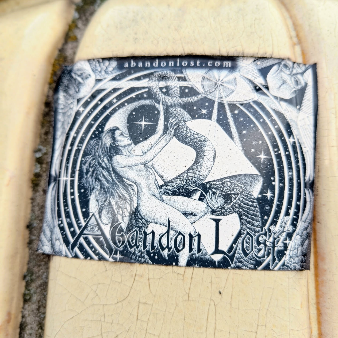 Black and white sticker with intricate artwork depicting a nude woman interacting with a large snake, all set against a backdrop of stars, celestial bodies, and swirling patterns. The sticker has the website abandonlost.com printed on the top. The text "Abandon Lost" is prominently featured at the bottom of the artwork.