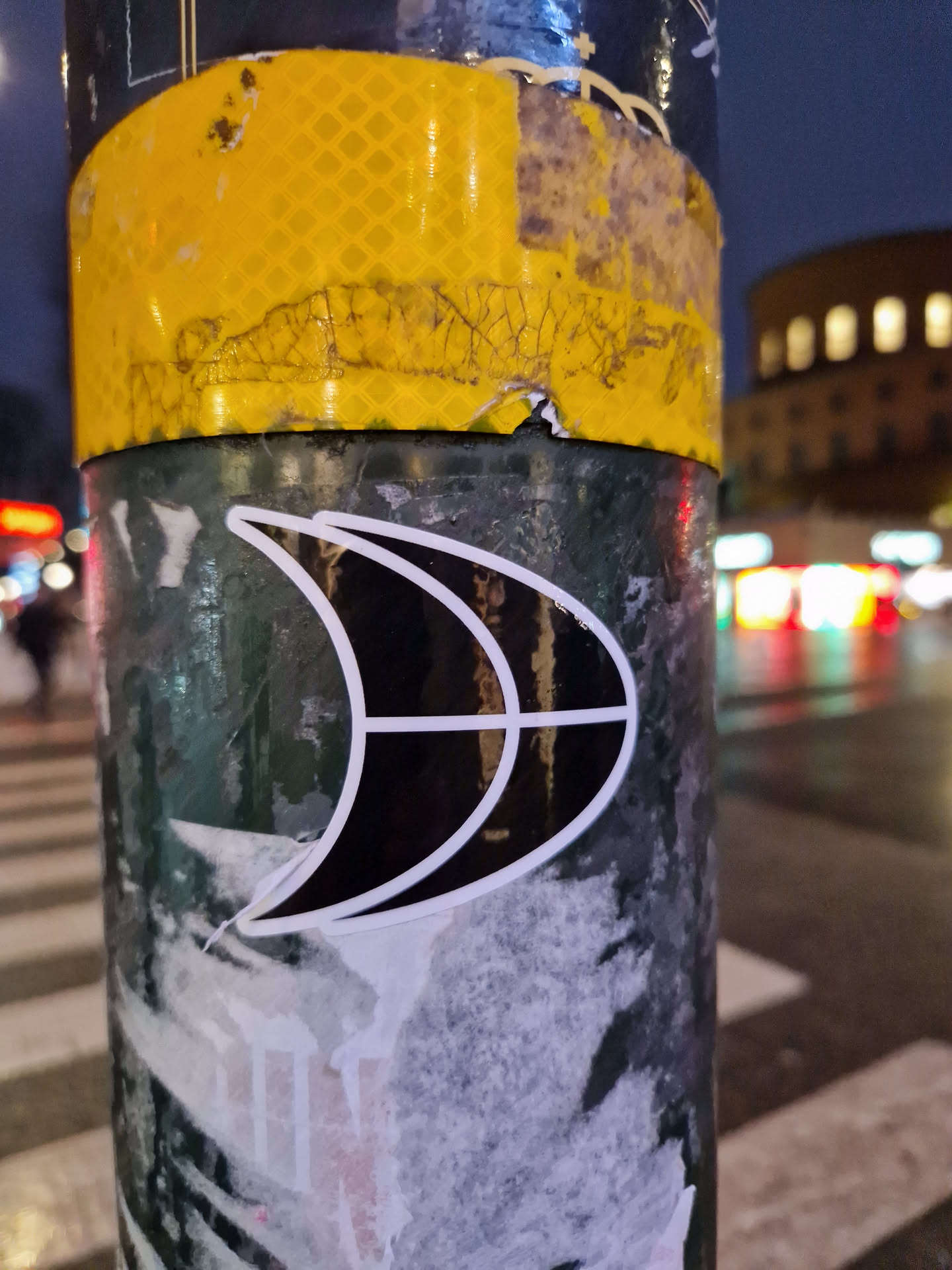 A close-up image of a black and white sticker affixed to a weathered pole at night. The sticker features a stylized crescent moon or abstract shape with internal lines, creating a simple yet eye-catching design.  The pole shows signs of wear, with other layers of stickers and markings visible beneath. The background is blurred, highlighting city lights at night.