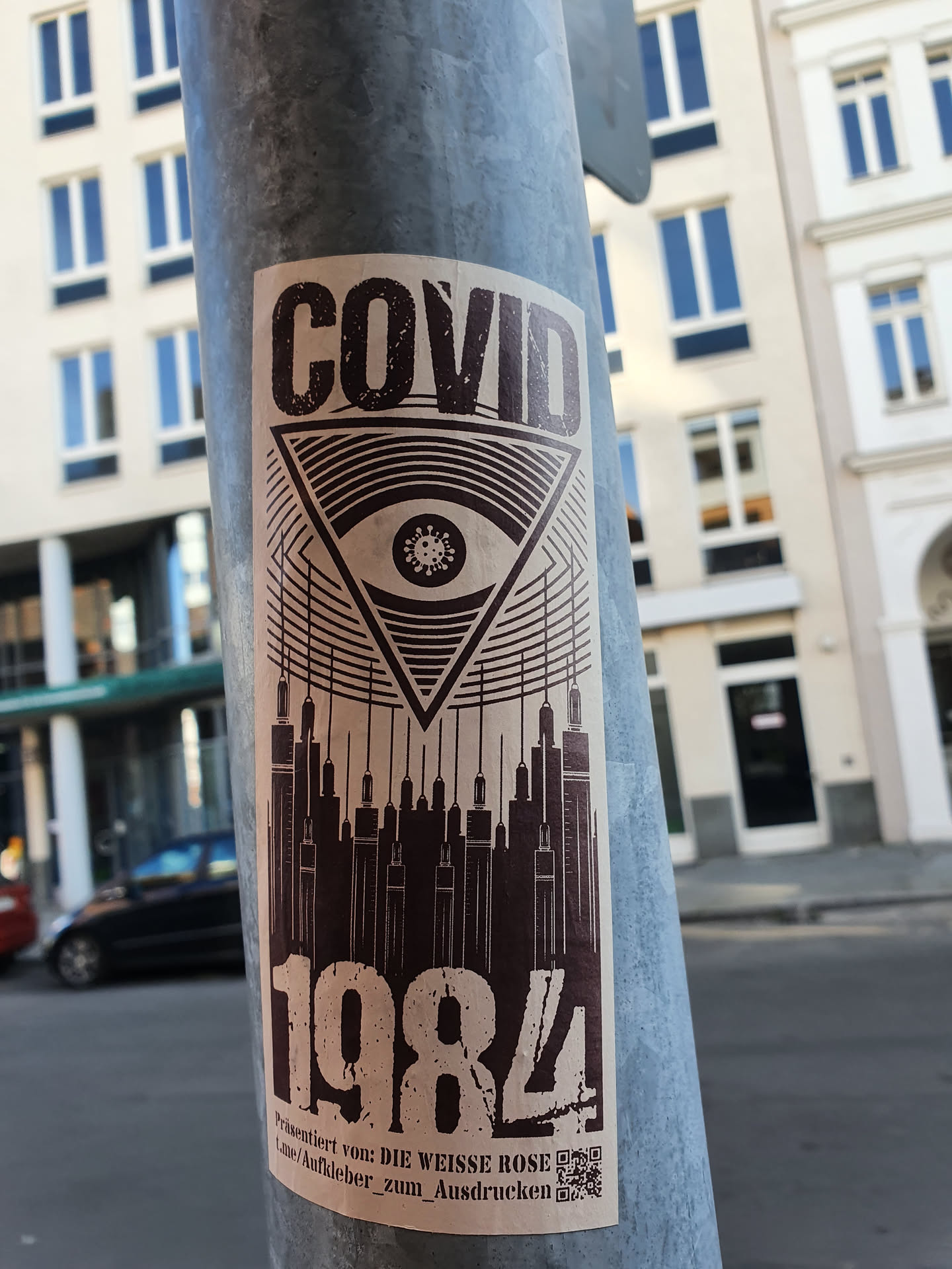 A sticker with a design combining imagery of COVID-19, an eye inside a triangle, syringes forming a cityscape, and the number 1984.  The text at the bottom indicates it's from Die Weisse Rose and provides a QR code. The overall aesthetic is dark and slightly dystopian.