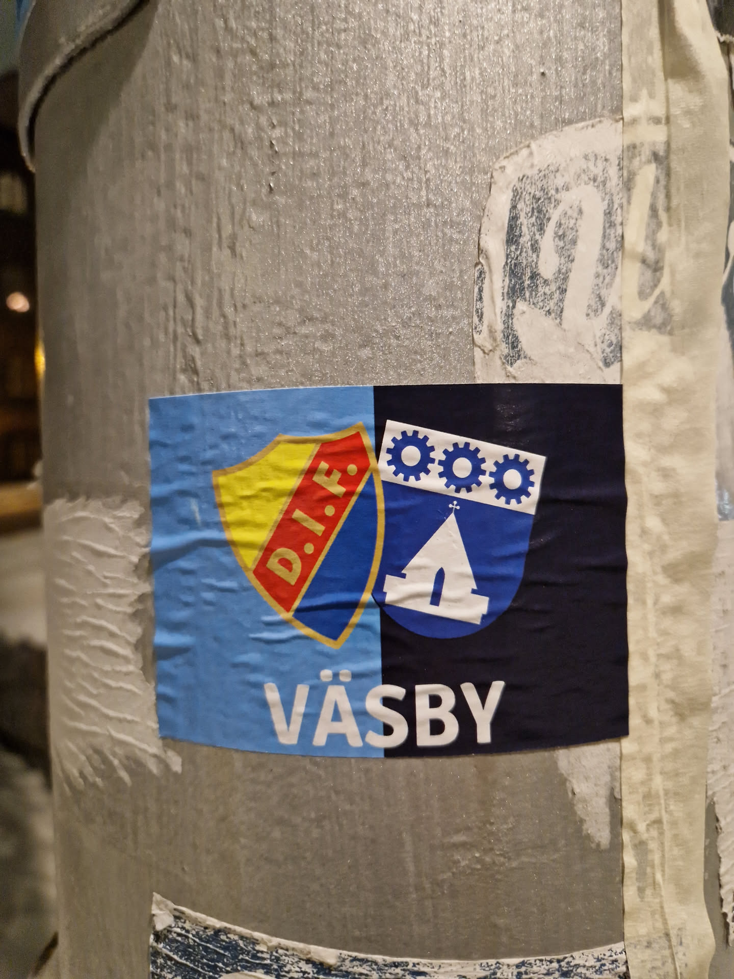A sticker featuring the emblem of Växjö. The sticker has a light blue background and contains two emblems, one with a red, yellow and blue shield and the other with a blue shield with a church and gears. The word "VÄSBY" is written in white letters below the emblems.