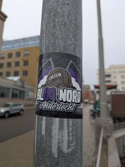 Street sticker A rectangular sticker adhering to a metal lamppost, associated with RSC Anderlecht football fans. At the center is an illustration of a classic brown leather laced football labeled '1908'. Behind the ball are two crossed flagpoles bearing purple and white striped flags. Below the ball, large stylized typography reads 'BLOC NORD', with 'BL' and 'NORD' in white and 'OC' in purple. Underneath, 'Anderlecht' is written in cursive script with a white fill and black outline.