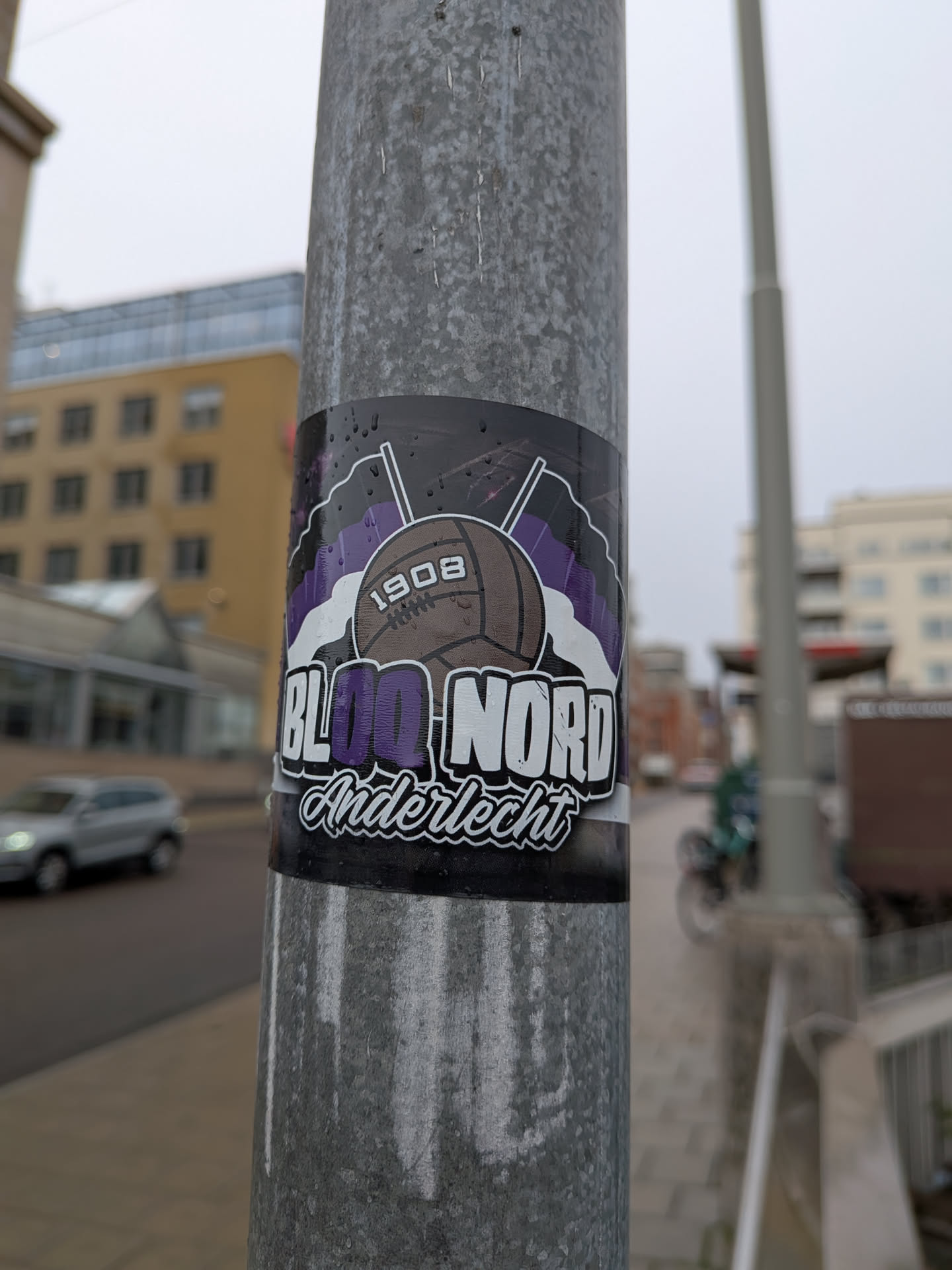A rectangular sticker adhering to a metal lamppost, associated with RSC Anderlecht football fans. At the center is an illustration of a classic brown leather laced football labeled '1908'. Behind the ball are two crossed flagpoles bearing purple and white striped flags. Below the ball, large stylized typography reads 'BLOC NORD', with 'BL' and 'NORD' in white and 'OC' in purple. Underneath, 'Anderlecht' is written in cursive script with a white fill and black outline.