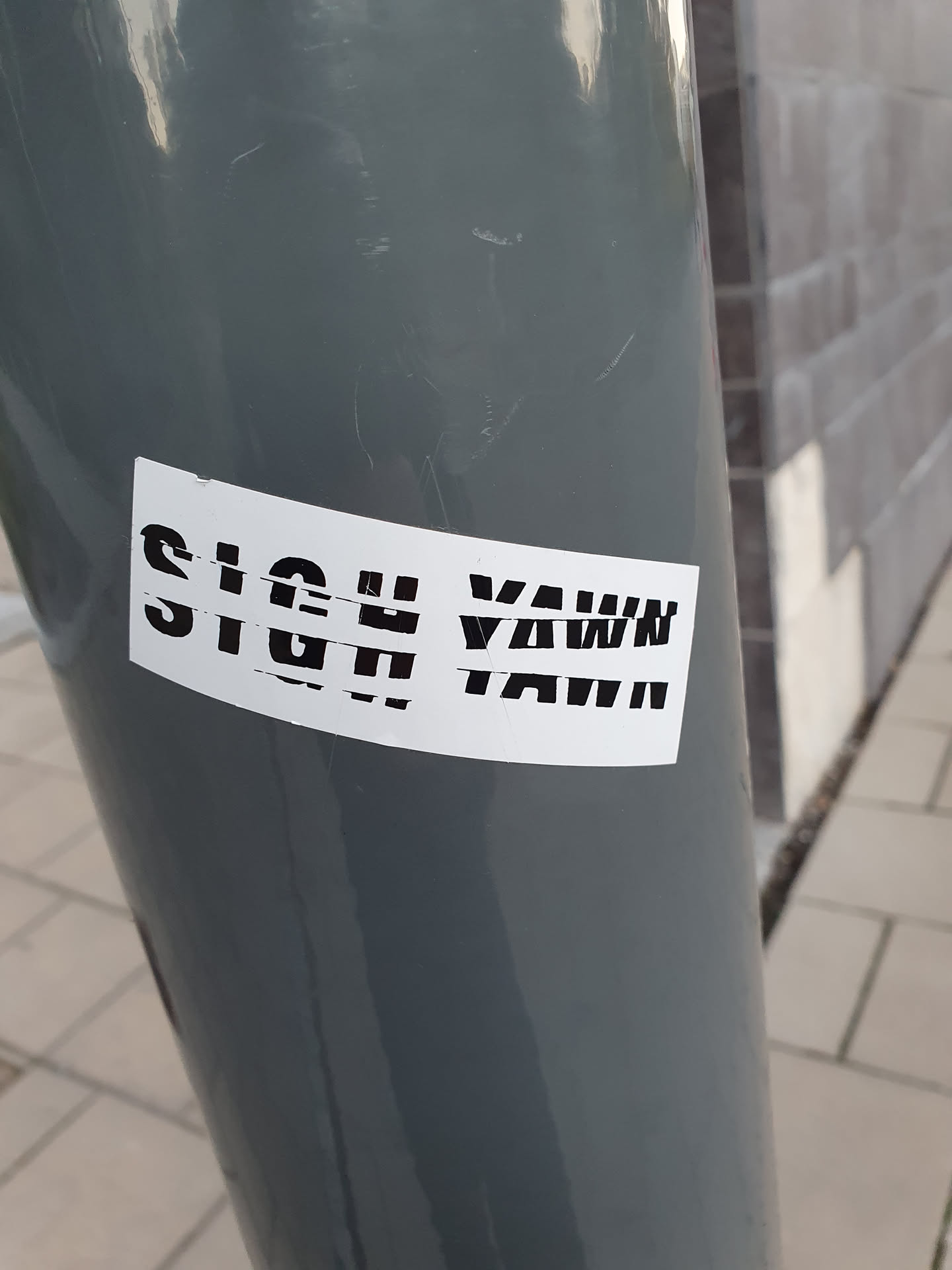 A white sticker with black text that says "SIGH YAWN" is affixed to a gray pole. The sticker is slightly damaged.