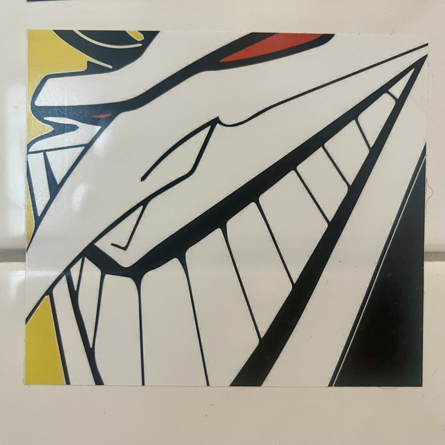 A square sticker showcasing an extreme close-up of a stylized character wearing a mask or featuring sharp, exaggerated facial contours. The character displays a massive, sinister grin with prominent square teeth, an elongated pointed nose or visor extending diagonally across the face, and a sliver of red near the eye. Bold black outlines define the shapes, with mostly white fill and small accents of bright yellow and red.