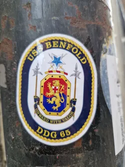 A sticker featuring the emblem of the USS Benfold (DDG-65). The emblem includes the ship's name, motto ("Onward With Valor"), and hull number.  The design is circular with a rope border and incorporates a coat of arms featuring a lion.