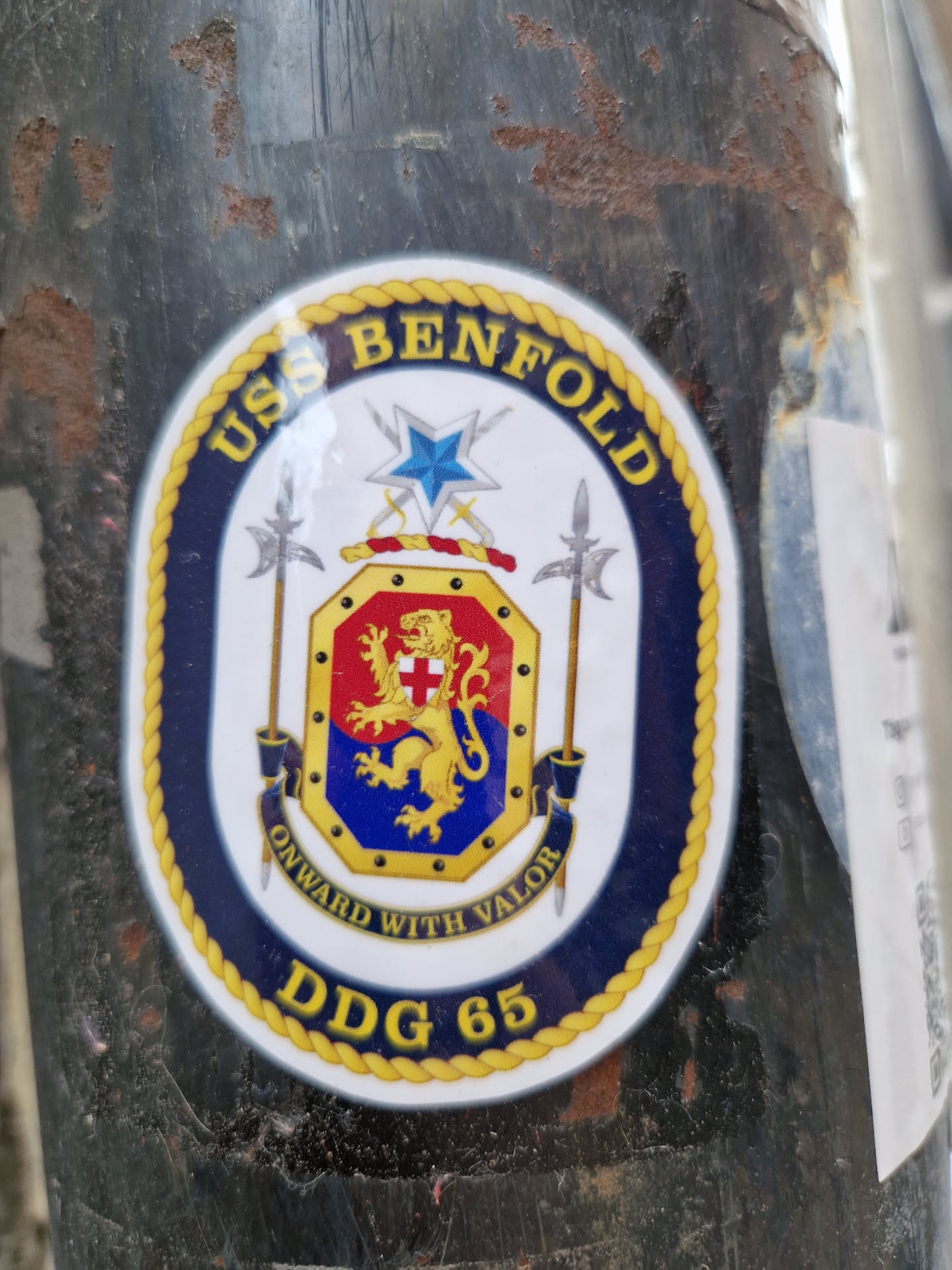 A sticker featuring the emblem of the USS Benfold (DDG-65). The emblem includes the ship's name, motto ("Onward With Valor"), and hull number.  The design is circular with a rope border and incorporates a coat of arms featuring a lion.