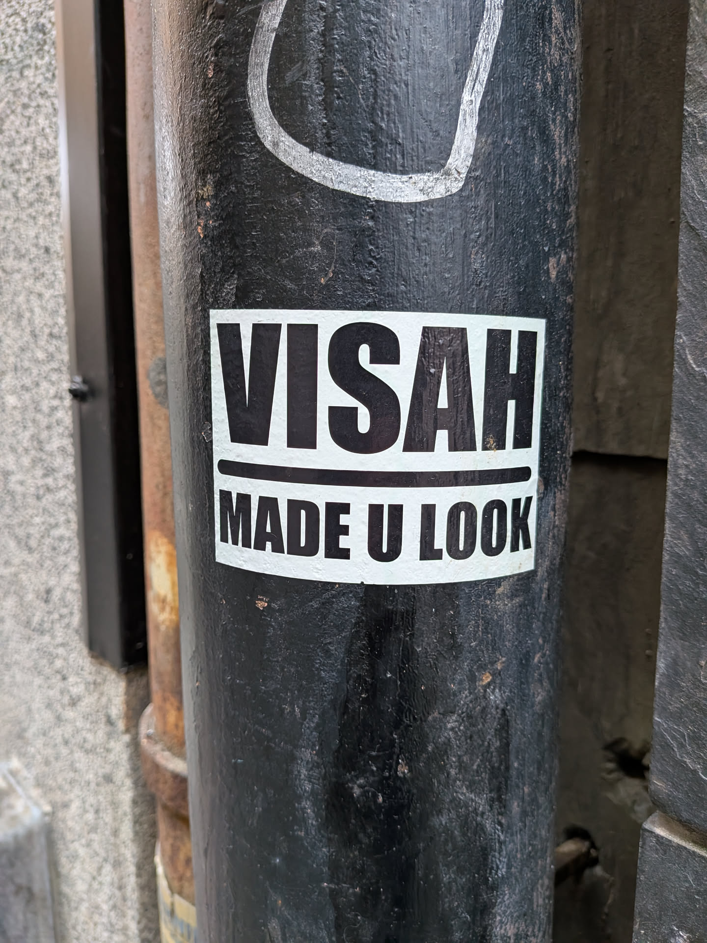 The sticker is rectangular with a white background and black text. The top line of text reads 'VISAH' in large, bold, sans-serif capital letters. Below this, separated by a thin black line, is the text 'MADE U LOOK', also in bold, sans-serif capital letters. The sticker appears to be applied to a black, cylindrical object, possibly a pole, which shows signs of wear and tear.