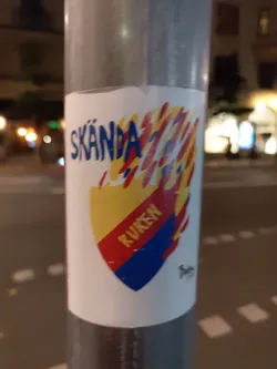 A sticker with a shield-like design featuring the words "Skända" and "Kuken". The shield is surrounded by flames, and the color scheme includes yellow, red, and blue. The style is bold and graphic.