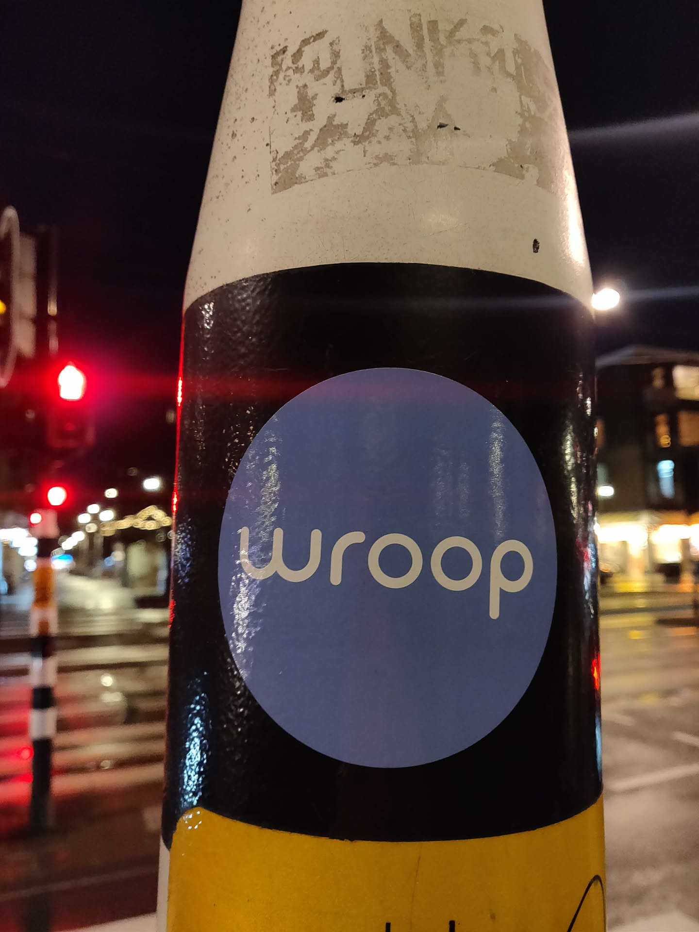 A round sticker with the word "wroop" in a modern font. The sticker is light purple with a white outline. It's placed on a black background, which in turn is on a yellow pole. The background is blurry, with city lights visible.