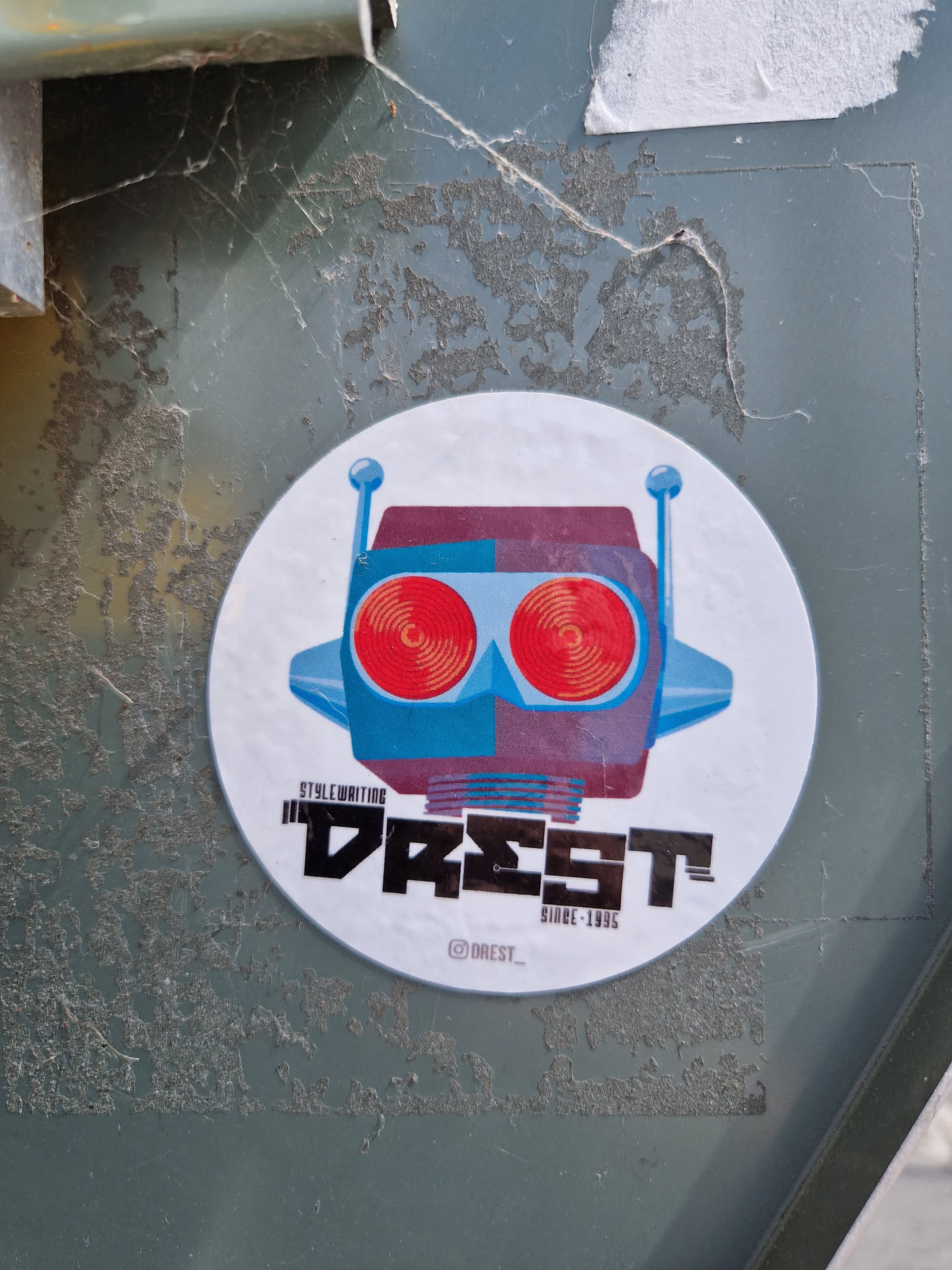 Round sticker featuring a stylized robot head with red eyes. The text "DREST" is prominently displayed below the robot, along with "Style Writing" and "Since 1995". The sticker has a minimalist design with a color palette of blues, reds, and purples.