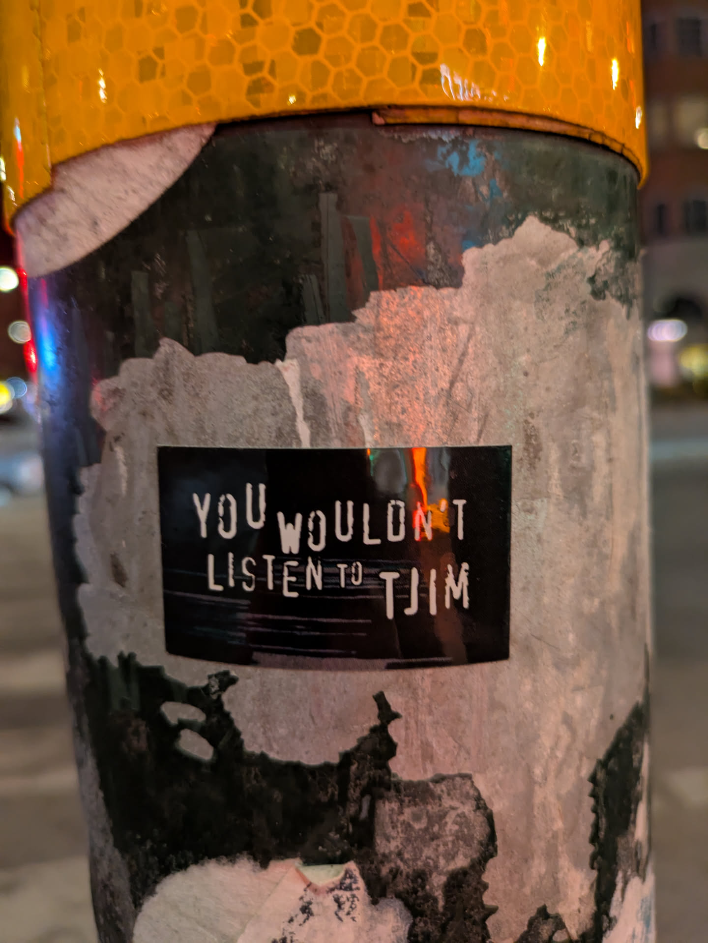 The sticker is rectangular with black background and white text that reads 'You wouldn't listen to TJIM'. It's attached to a weathered light pole with remnants of old adhesives and torn stickers.
