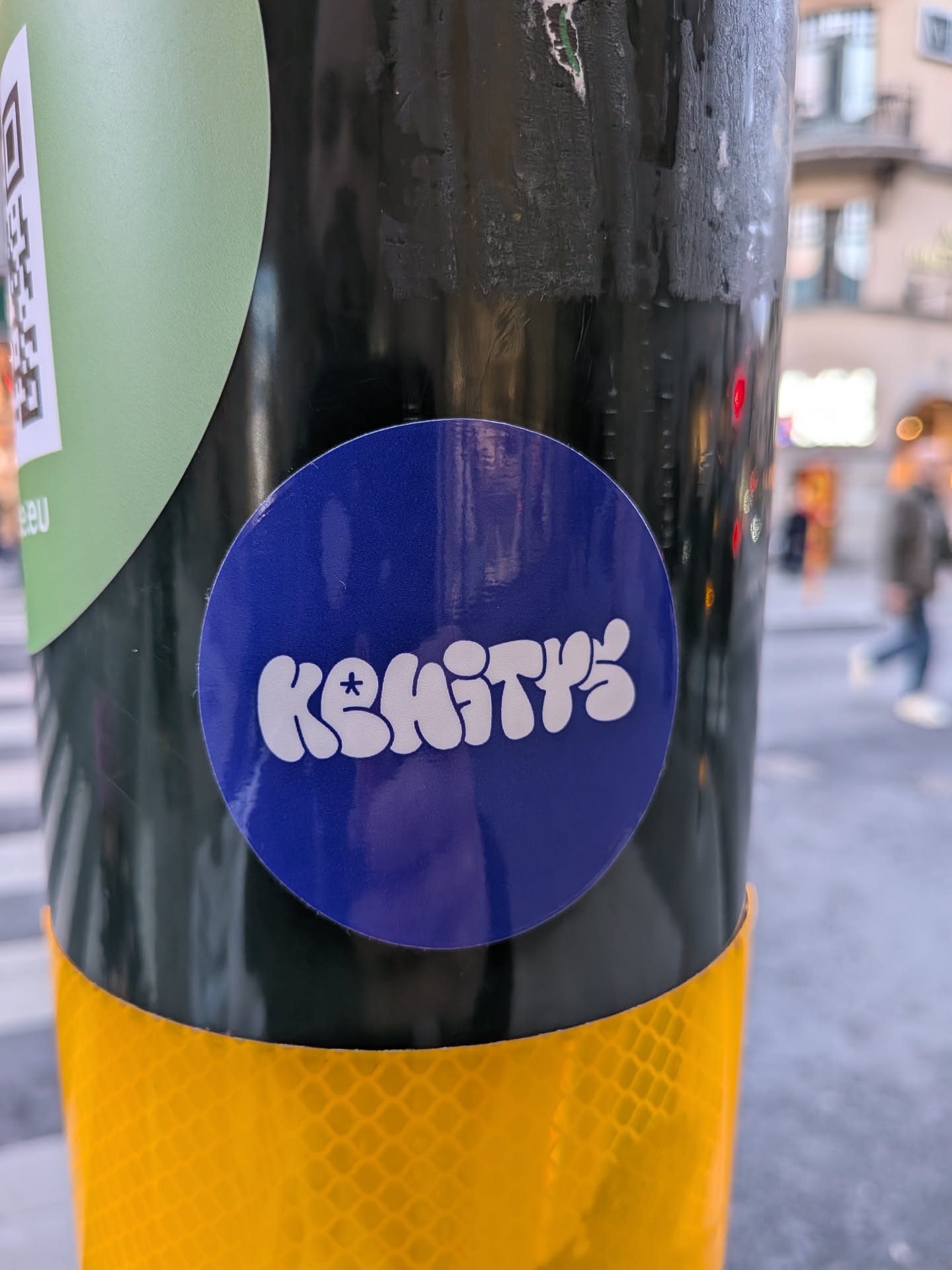 A circular, deep blue sticker featuring the word 'KEMITYS' in bold, white, bubble-style letters. The 'E' character uniquely incorporates a small asterisk-like symbol within its upper loop. The letters have a soft, rounded appearance. The sticker itself has a slight sheen, indicating a glossy finish, and is adhered to a dark, textured pole, positioned just above a yellow reflective strip.
