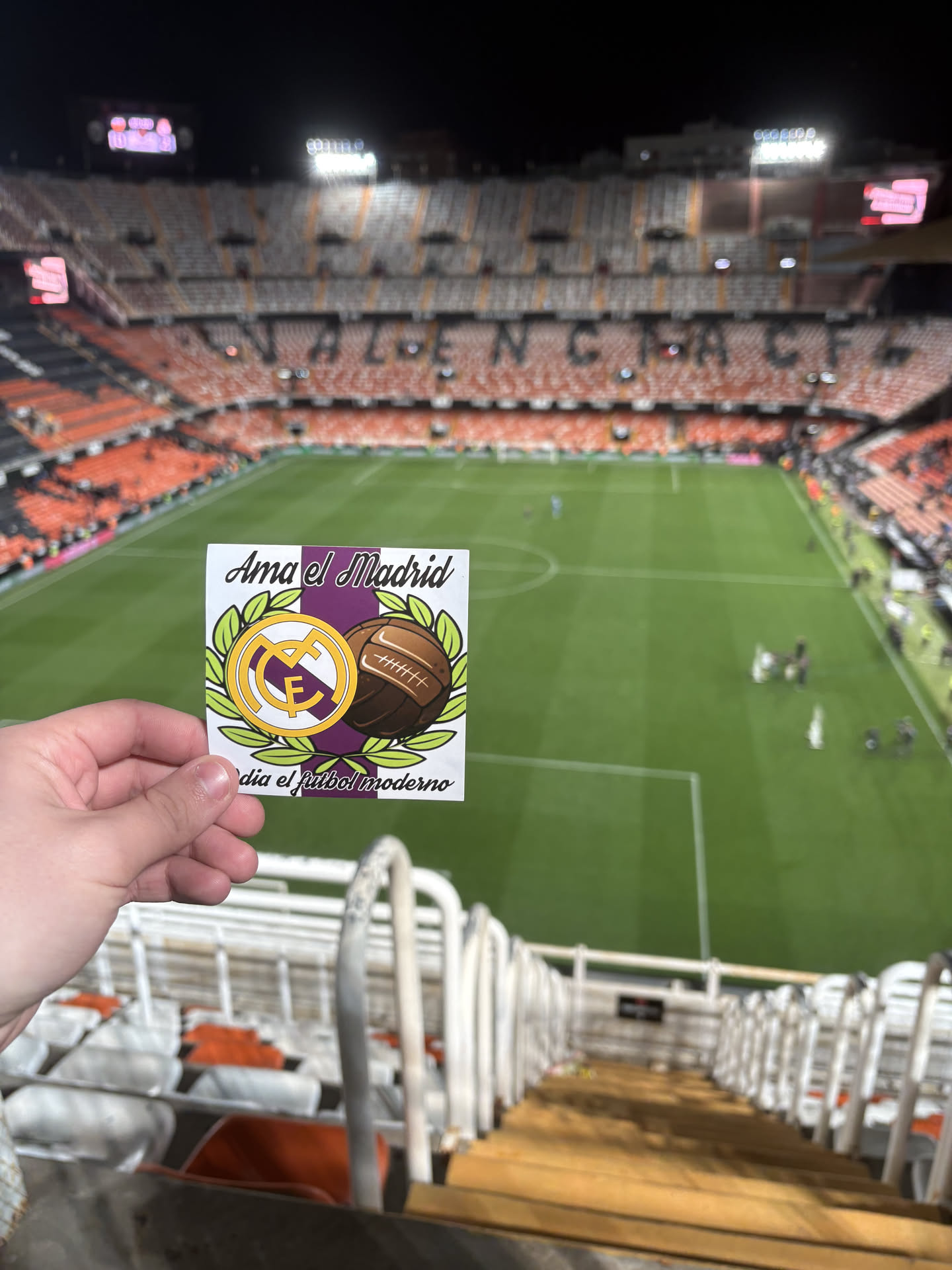 The sticker is a square design with a white border. The central design features a vertical purple stripe running behind two main elements: on the left, the iconic gold and white Real Madrid C.F. logo (a stylized 'MCF' with a crown) set against a purple circle, and on the right, a vintage-style brown leather football with visible lacing and stitching. Both the logo and the football are framed by a vibrant green laurel wreath. Above the central design, in a stylized black font, is the text 'Ama el Madrid', and below, 'odia el fútbol moderno'. The overall aesthetic combines club pride with a nostalgic view of the sport.