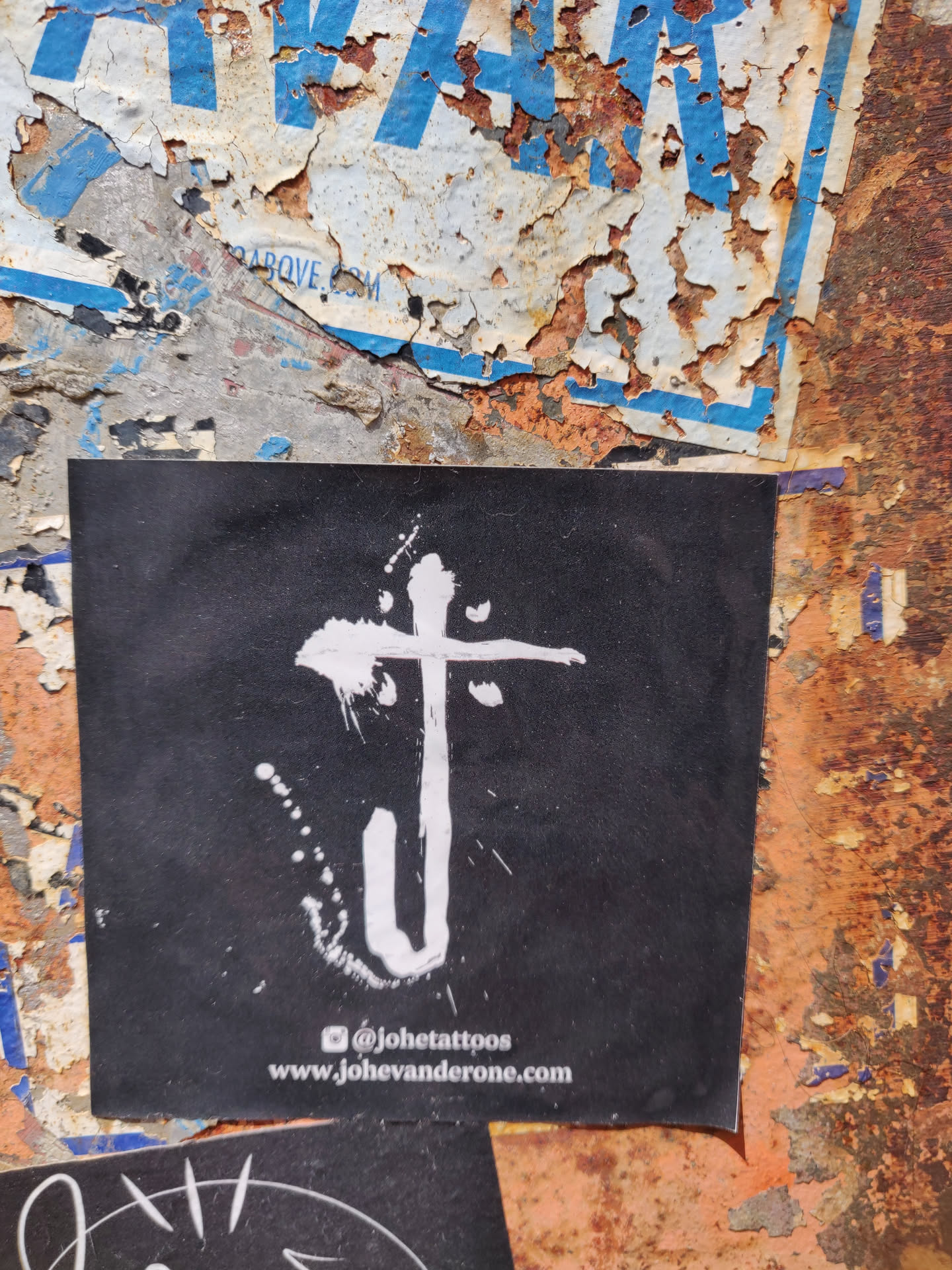 A black square sticker with a white abstract design in the center. At the bottom of the sticker, there is a social media handle and a website address. The sticker is on a rusty wall.