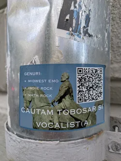 A blue sticker featuring an image of a man on a horse is adhered to a metallic pole. The sticker lists musical genres such as Midwest Emo, Indie Rock, and Math Rock. Below the image, text indicates the band is searching for a drummer and a vocalist. A QR code is located in the upper right corner of the sticker.