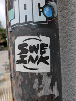 The sticker is square with slightly irregular edges, predominantly white with bold black lettering. The text reads 'SWE INK' in a thick, stylized font. A large, curved black line is positioned above the text, and a smaller, curved line is beneath it, resembling a smile. The sticker appears to be adhered to a dark, weathered pole, with peeling paint visible around its edges.