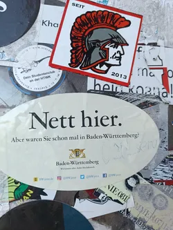 Oval-shaped sticker with a message in German, promoting tourism in Baden-Württemberg.  It includes the coat of arms of Baden-Württemberg and social media links.