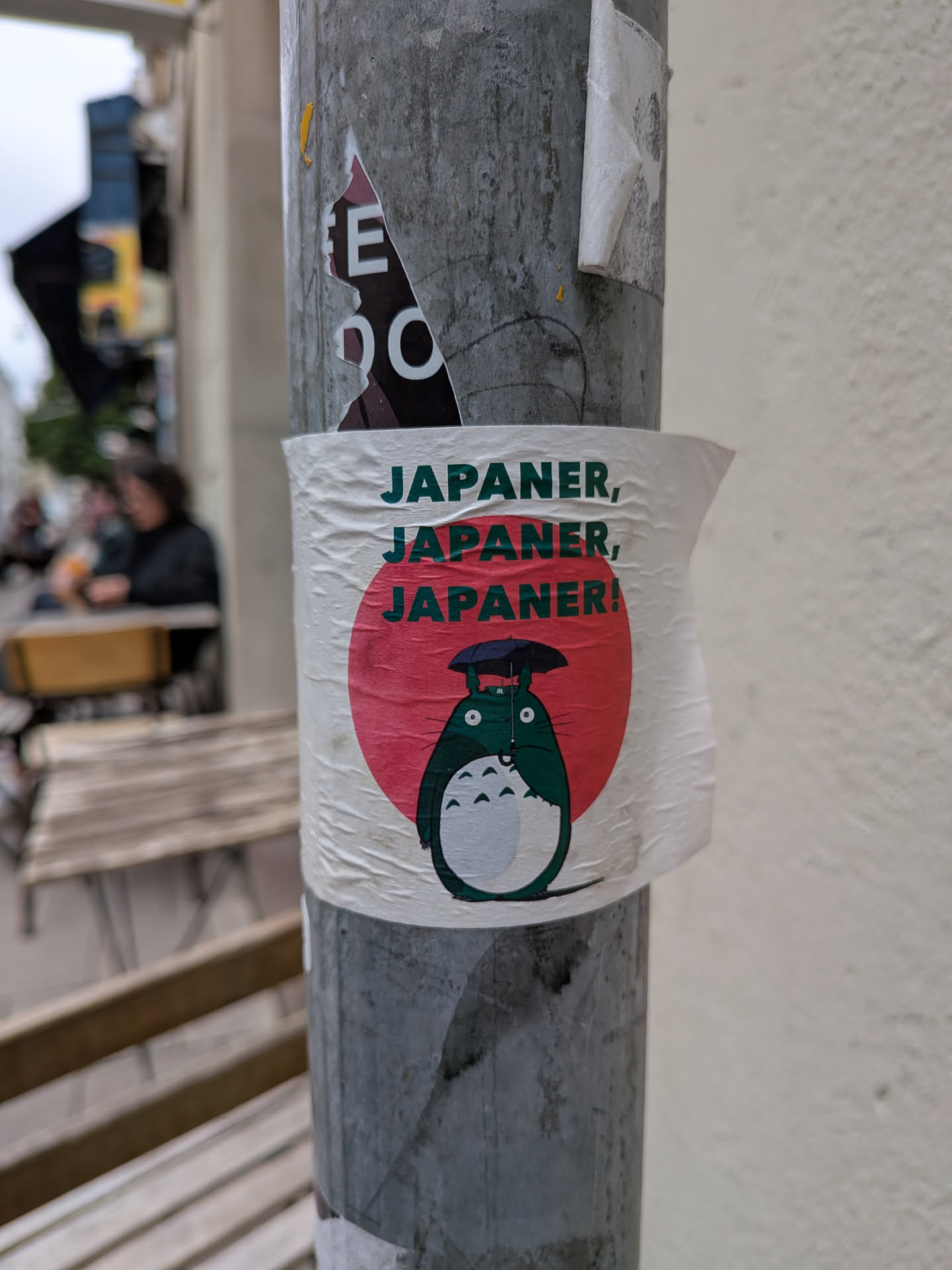 The sticker features a round, white background with a prominent red circle in the center, reminiscent of the Japanese flag. Below the red circle, the character Totoro from Studio Ghibli is depicted in green and white. Totoro is holding a black umbrella over its head. Above Totoro and the red circle, the text "JAPANER, JAPANER, JAPANER!" is printed in a dark green, bold font, arranged in three lines.