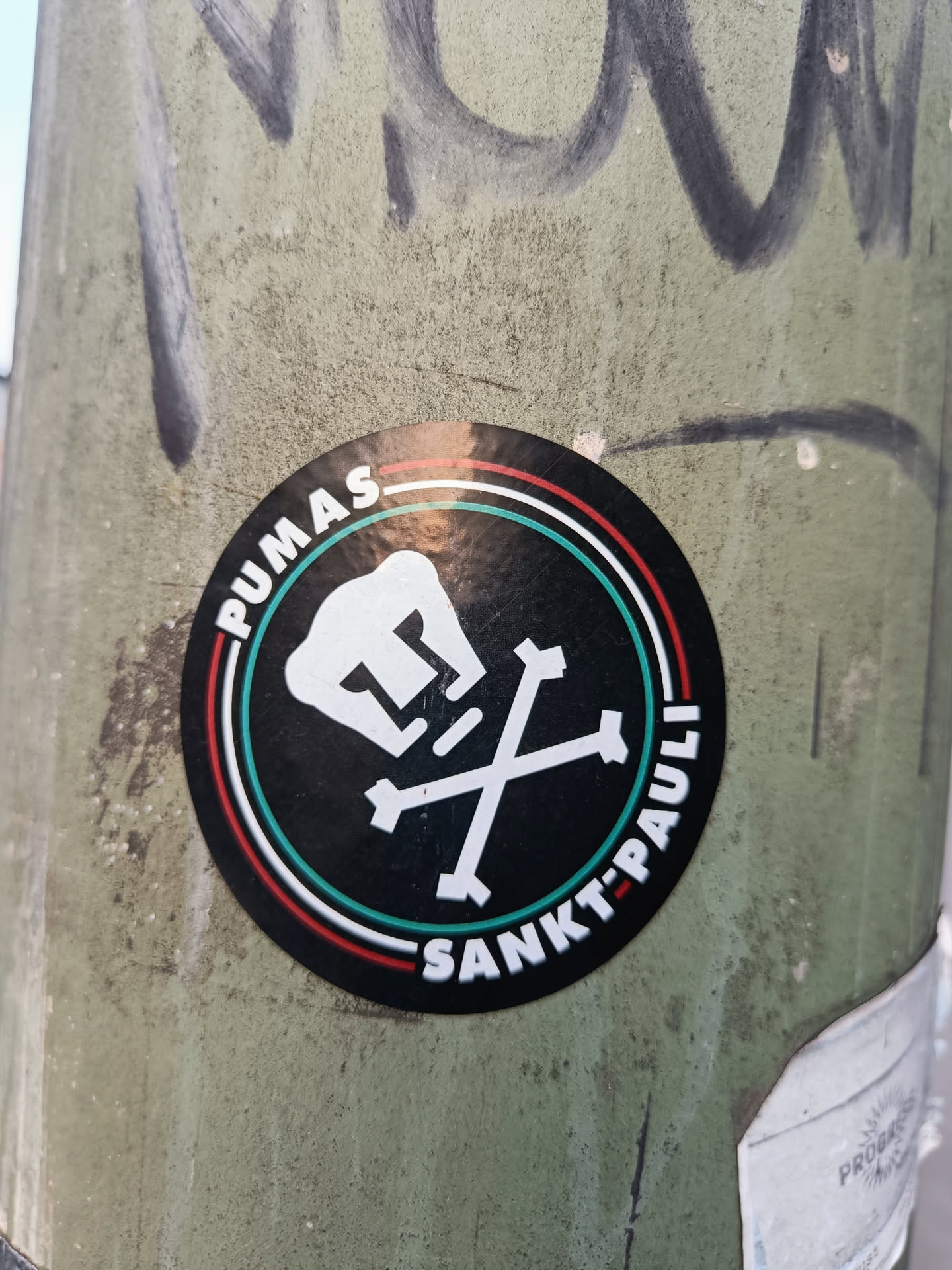 A round sticker featuring a Puma head and crossed bones, with the text "PUMAS" and "SANKT-PAULI" in a circular design. The color scheme is black with green, red and white accents.