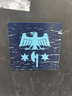 This sticker shows some wear and tear, giving it a vintage look. The design is simple, featuring a light blue eagle positioned above an axe with two stars on either side. The color scheme is primarily dark blue and light blue, giving it a somewhat muted look.