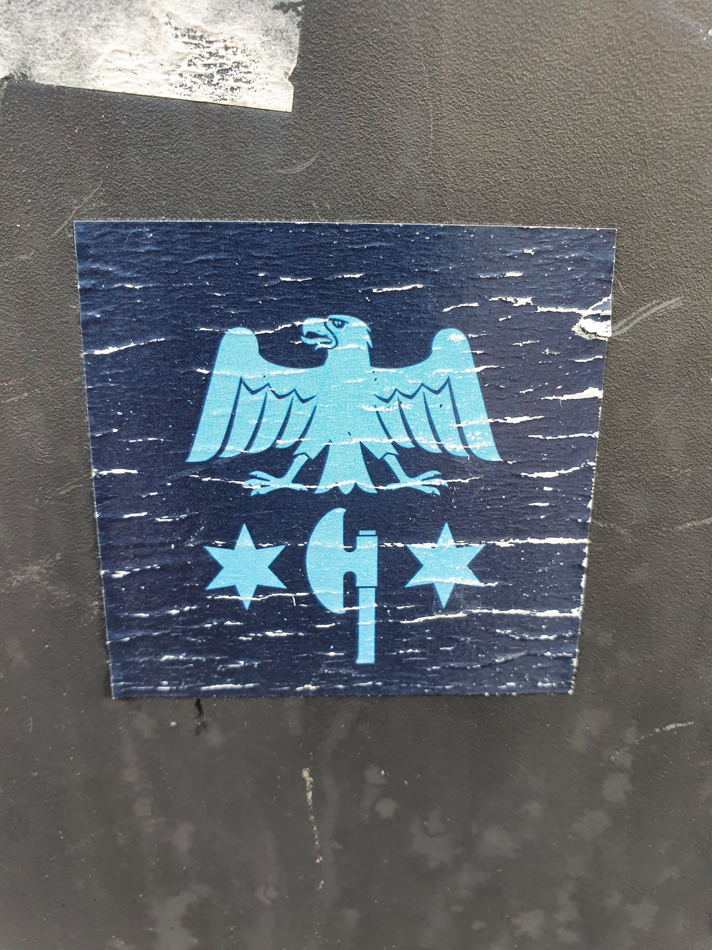 This sticker shows some wear and tear, giving it a vintage look. The design is simple, featuring a light blue eagle positioned above an axe with two stars on either side. The color scheme is primarily dark blue and light blue, giving it a somewhat muted look.