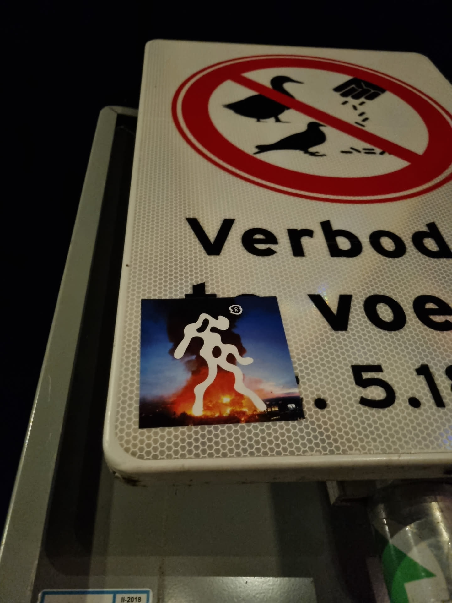 A sticker featuring a stylized figure set against a backdrop of fire and smoke. The sticker is affixed to a traffic sign that prohibits feeding birds. The figure's design is abstract and bold, suggesting themes of protest or rebellion. 